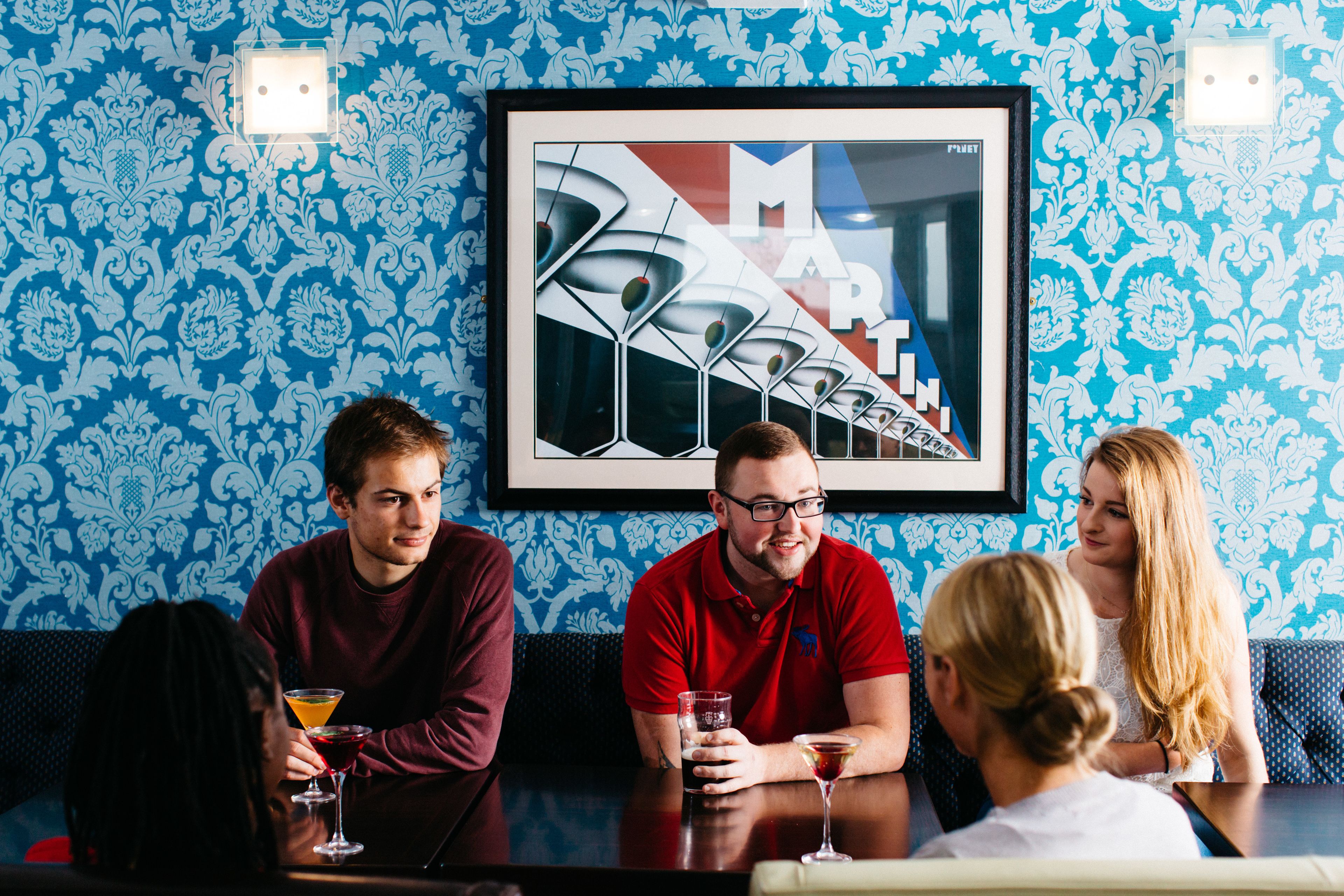 A group of people seated at a table in a room with patterned wallpaper and a framed artwork on the wall.