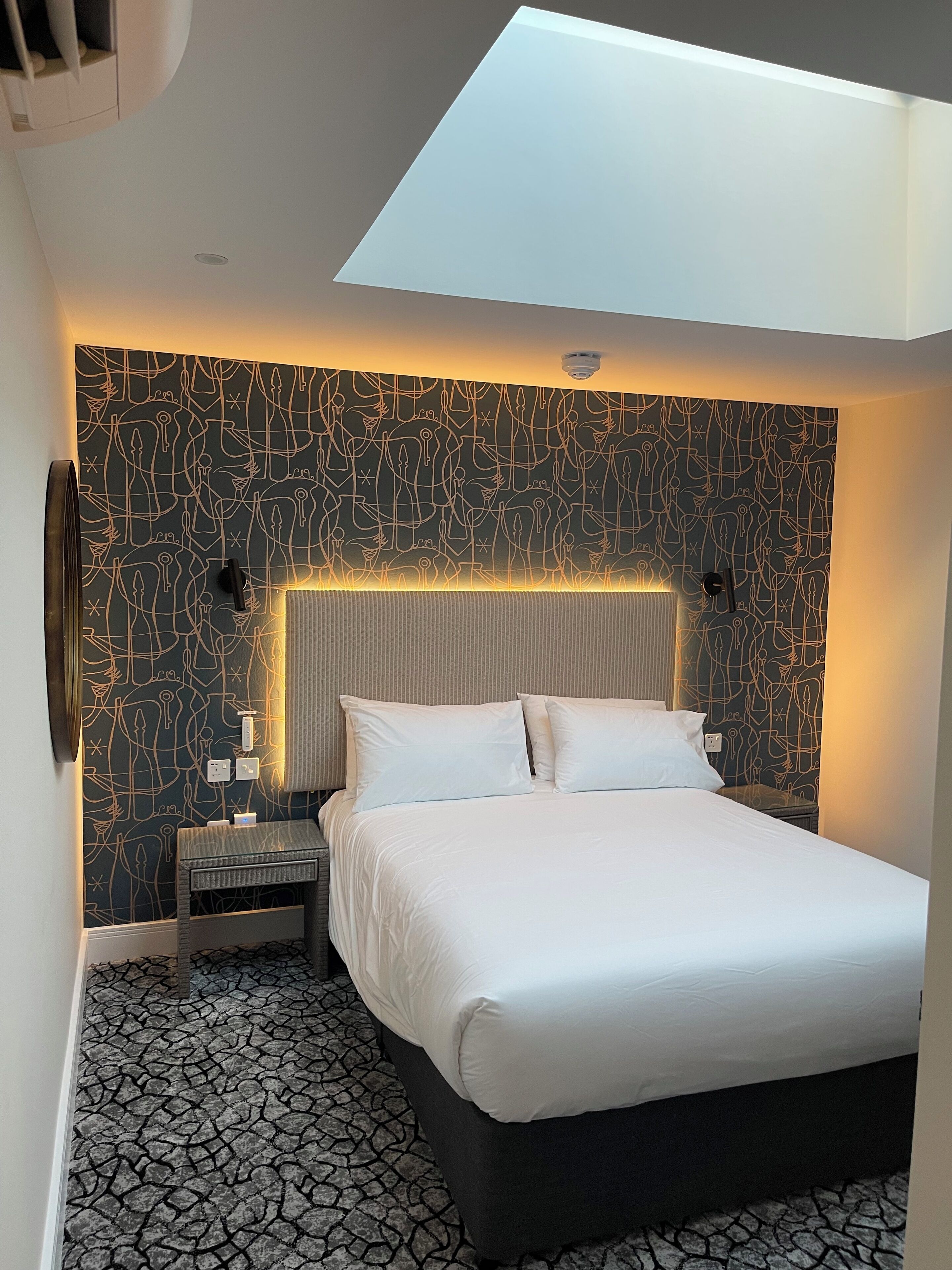 A modern hotel room with a skylight, a patterned wall, a large bed, and a nightstand with a lamp.