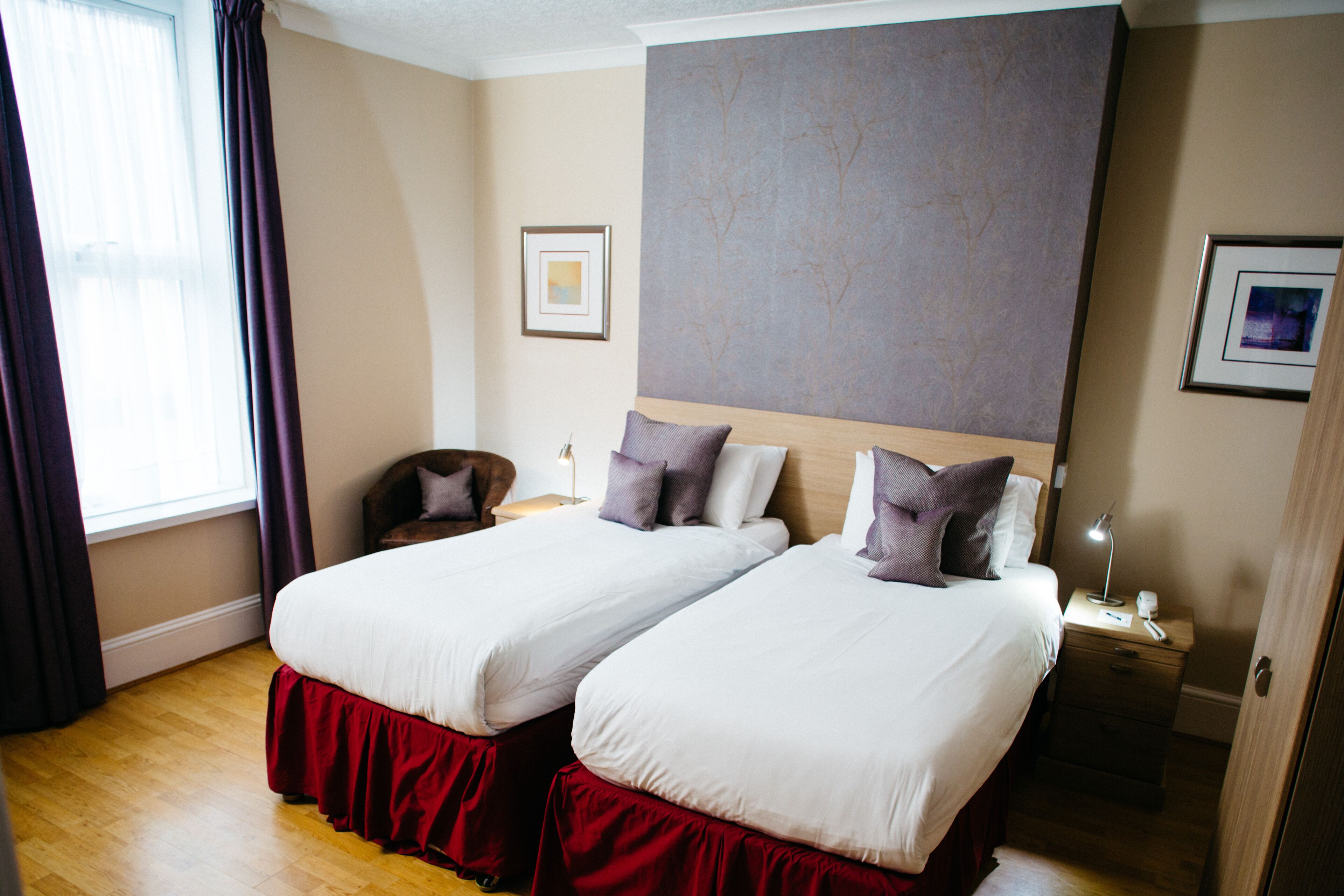 A hotel room with two single beds, a wooden headboard, a nightstand with a lamp, and a framed picture on the wall.