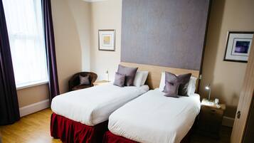 A hotel room with two single beds, a wooden headboard, a nightstand with a lamp, and a framed picture on the wall.