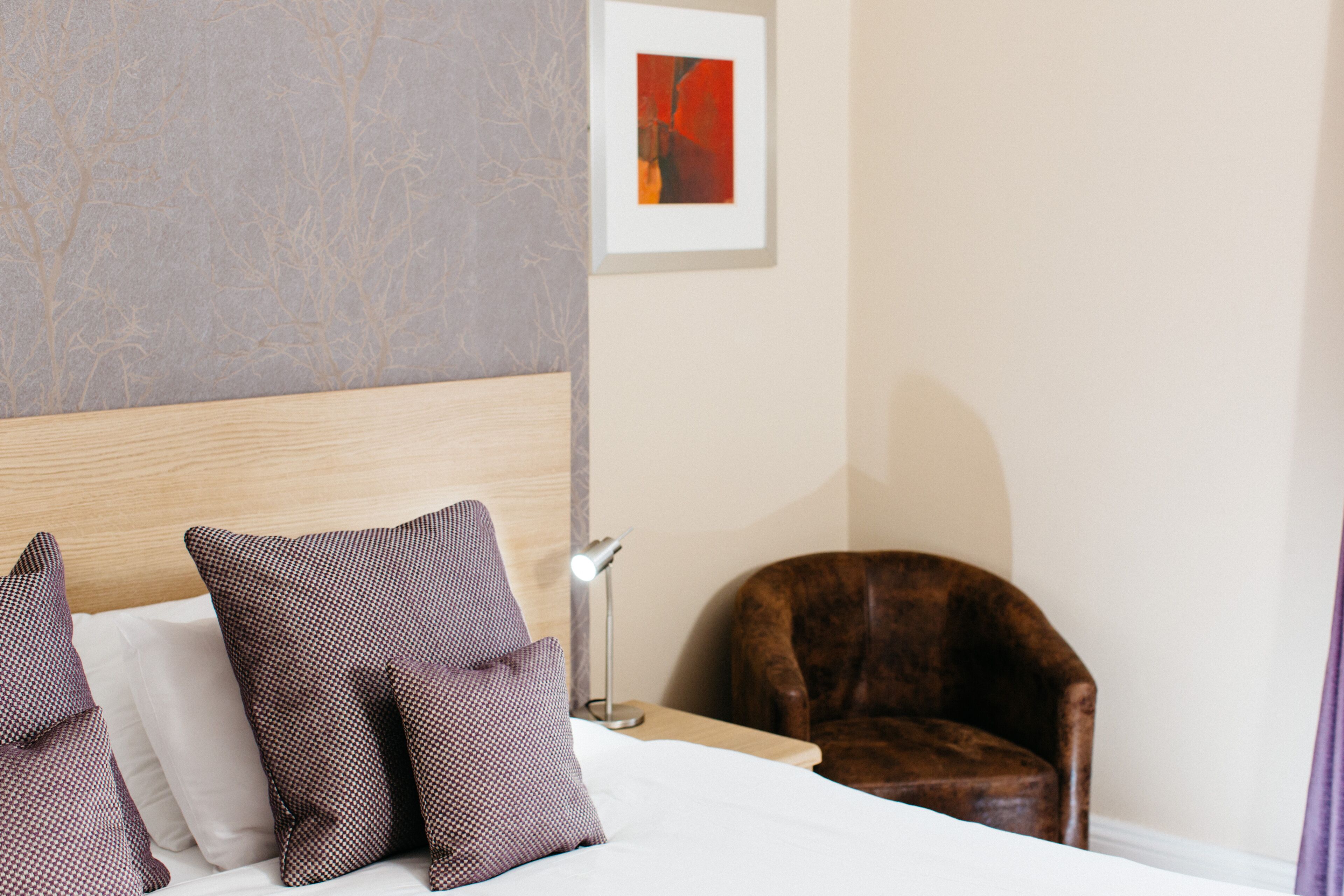 Une chambre d’hôtel avec une tête de lit en bois, un lit agrémenté d’oreillers à motifs, un fauteuil en cuir marron et une œuvre d’art encadrée au mur.