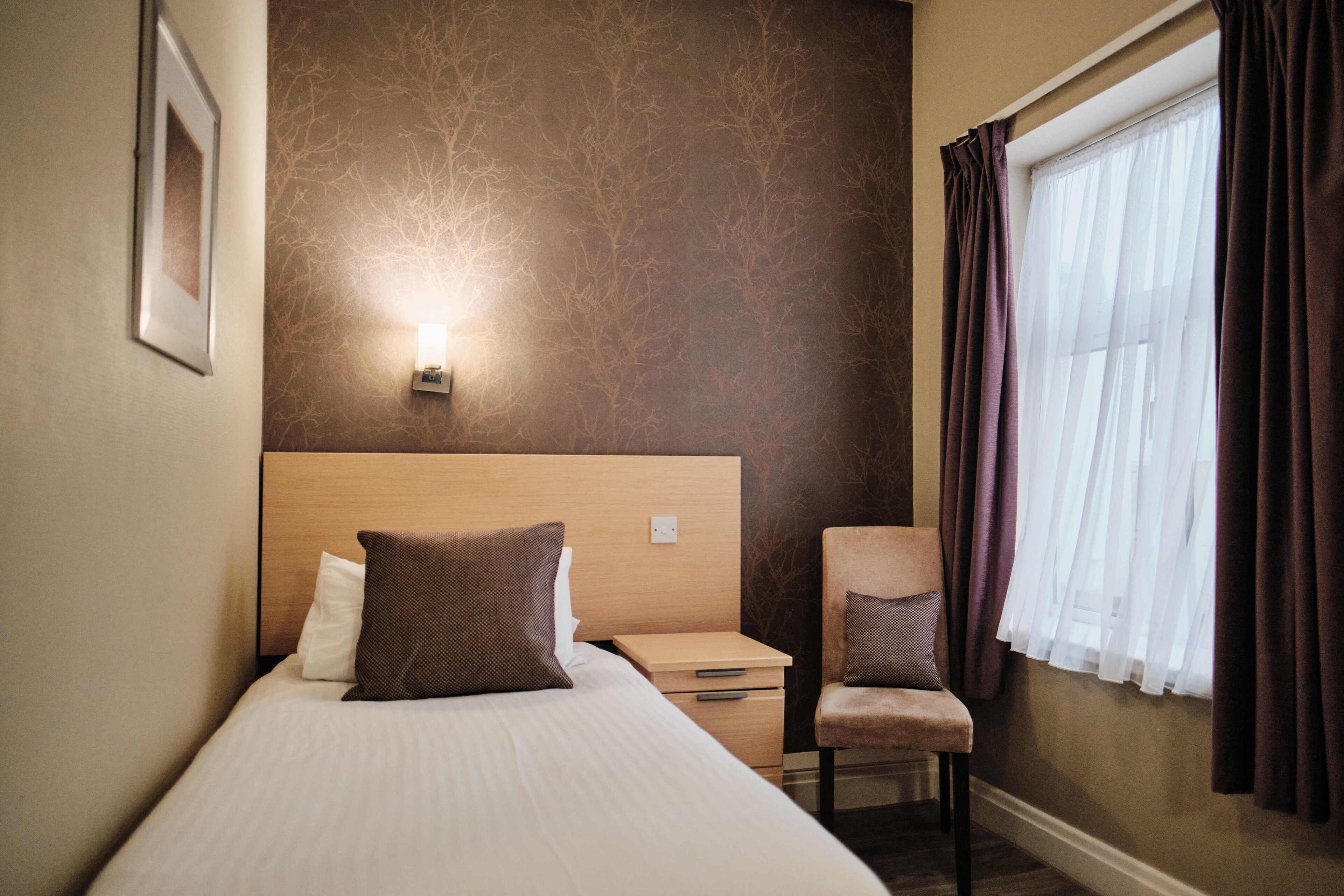 Habitación de hotel con cama, mesita de noche, una silla, una lámpara de pared y una ventana con cortinas.