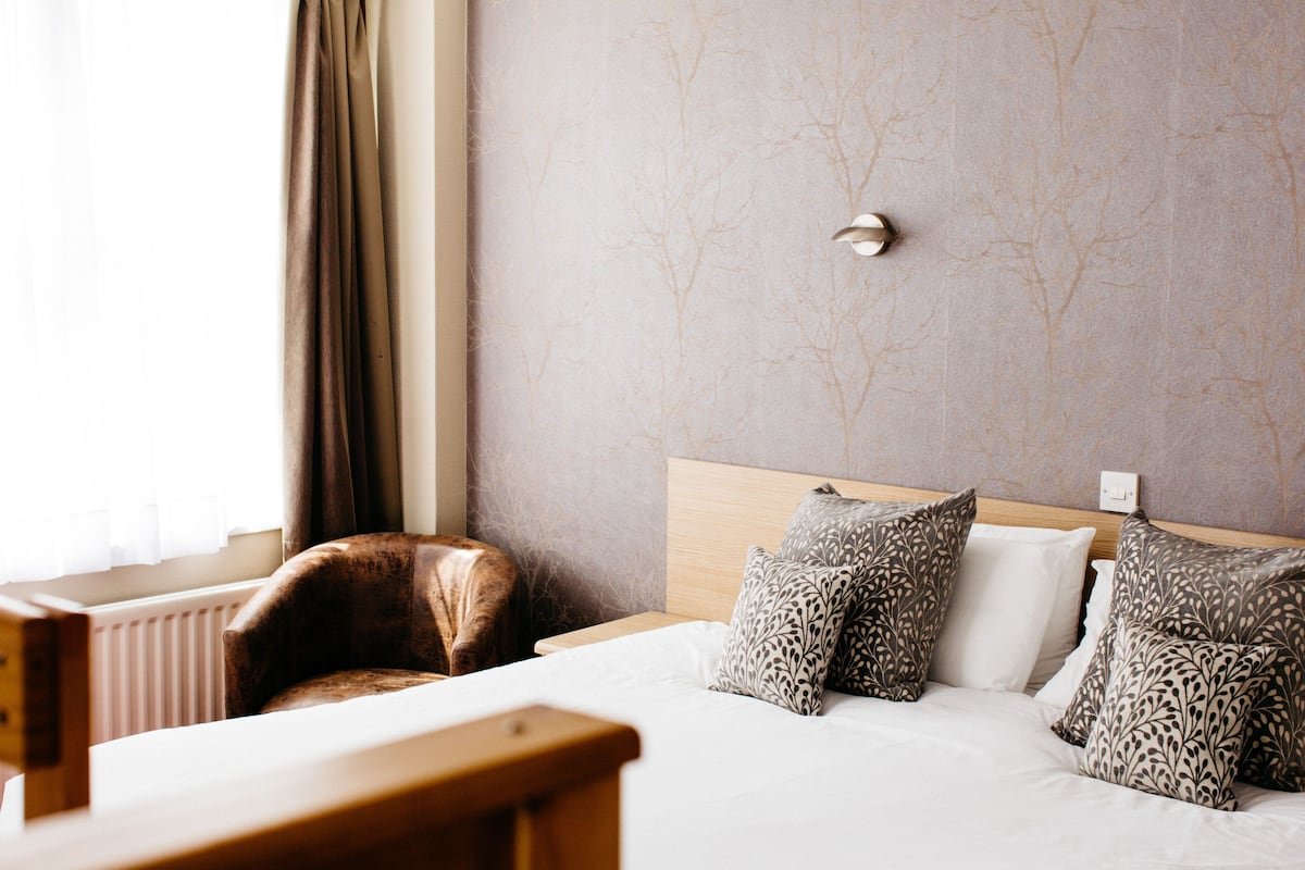 Habitación de hotel con cama, almohadas, cabecero de madera, radiador y ventana con cortinas.