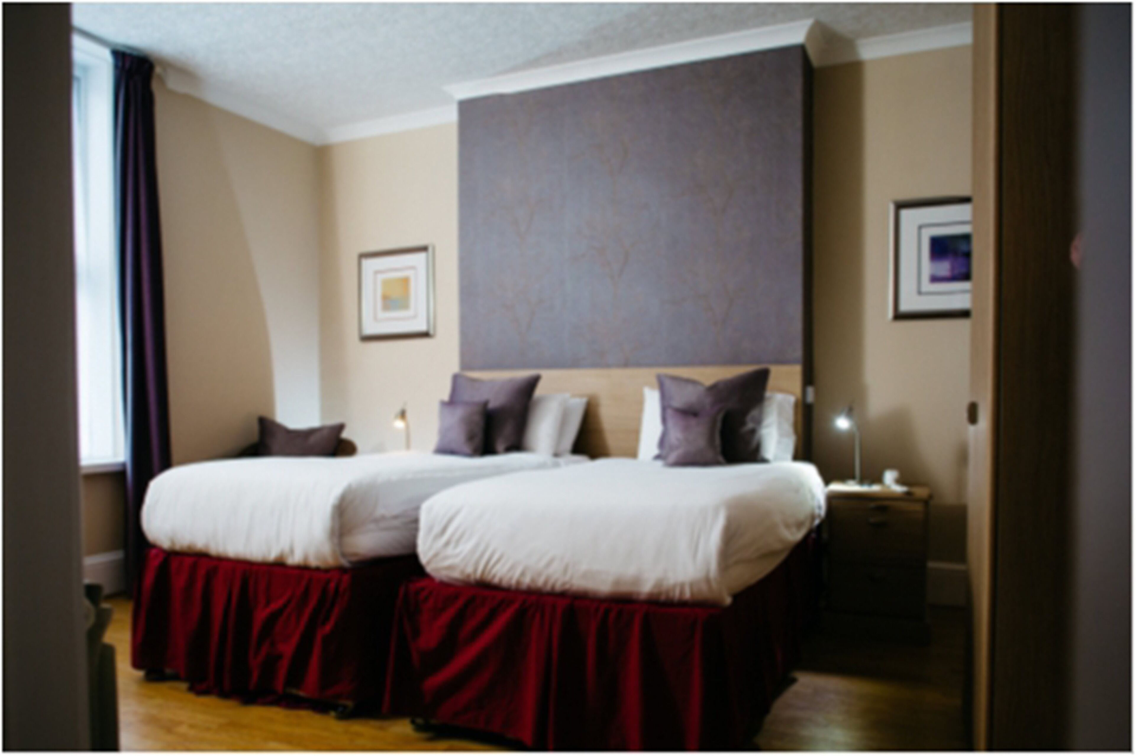 A double bed with a red skirt, two bedside tables, a nightstand, and a large window with curtains.