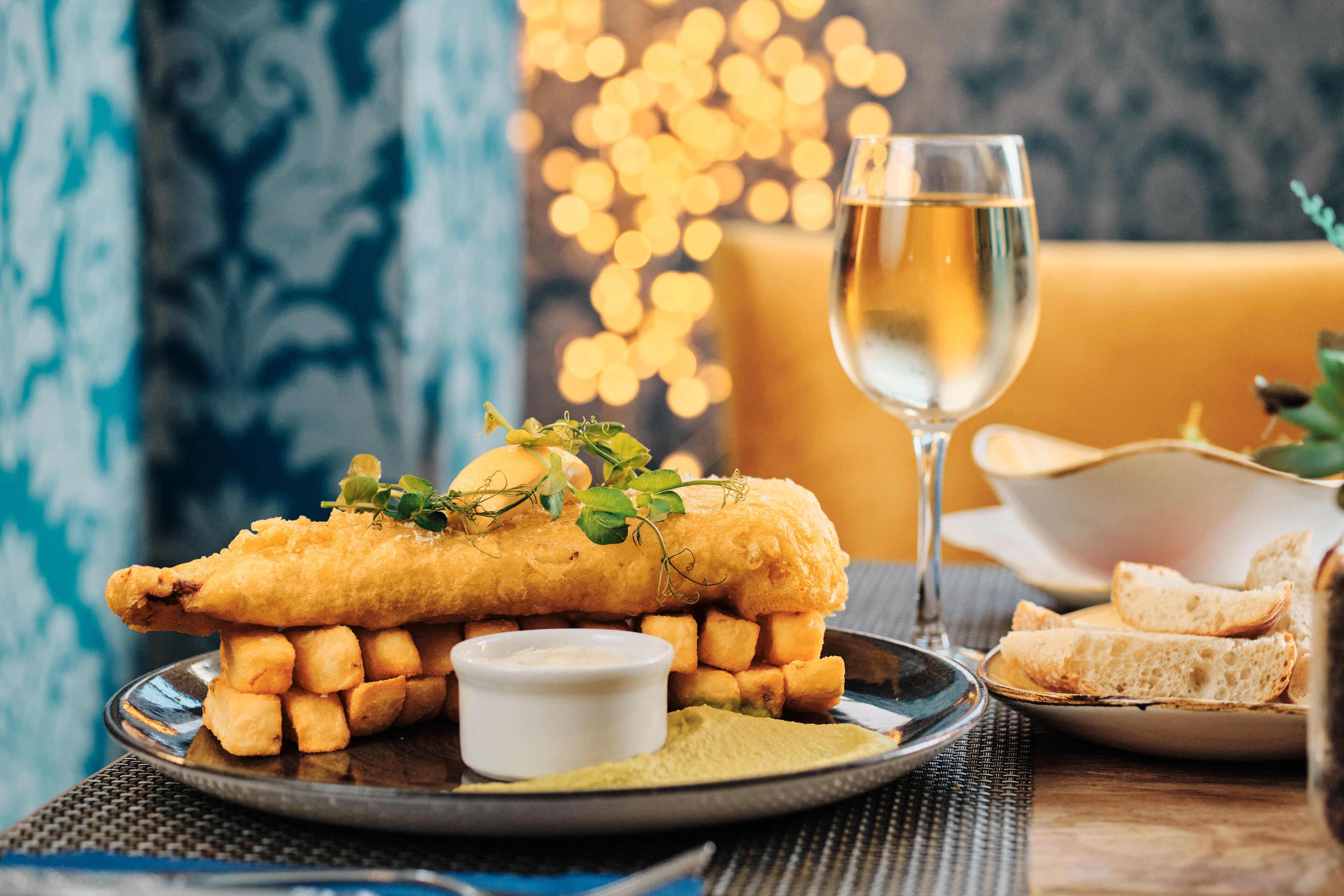 A plate of fish and chips with a glass of white wine, accompanied by a small bowl of tartar sauce and a side of fries.