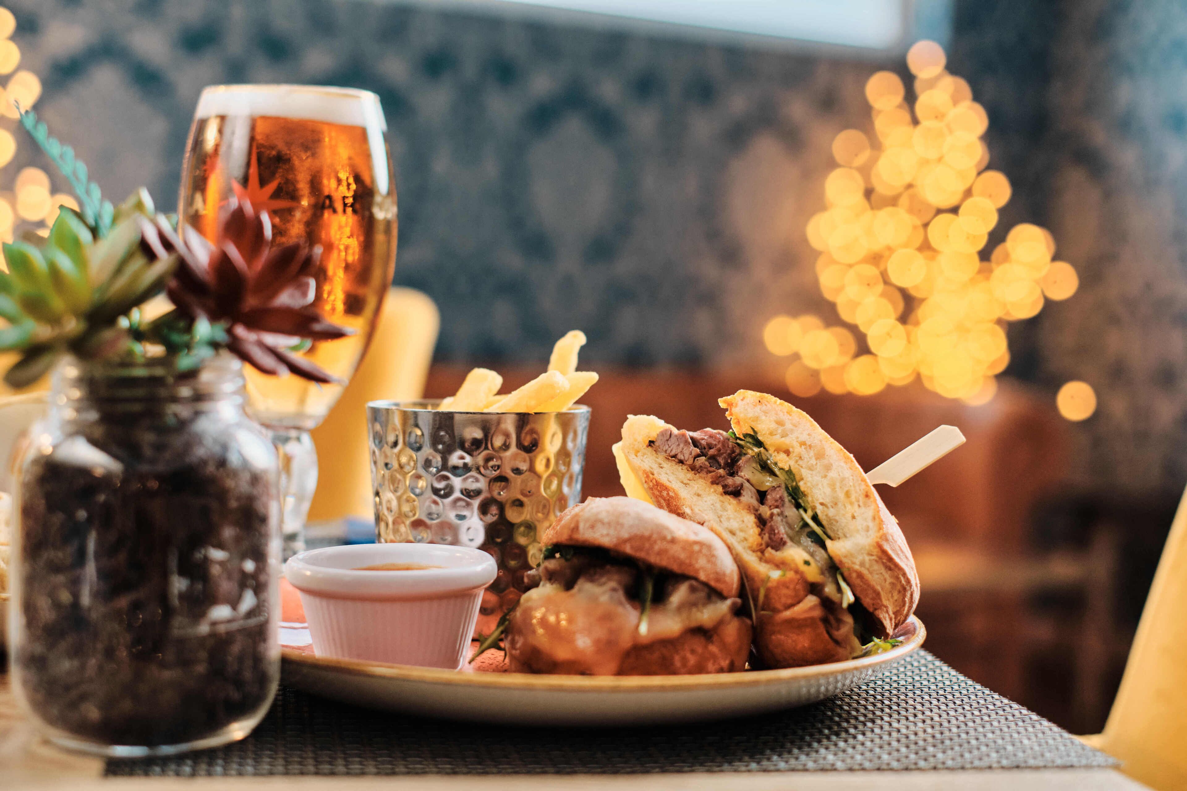 A plate of food including a sandwich and fries, a glass of beer, and a small bowl of sauce.