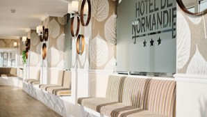 Zona de recepción de un hotel con un largo banco y una pared con diseño de hojas, y un cartel que dice "HOTEL DE NORMANDIE".
