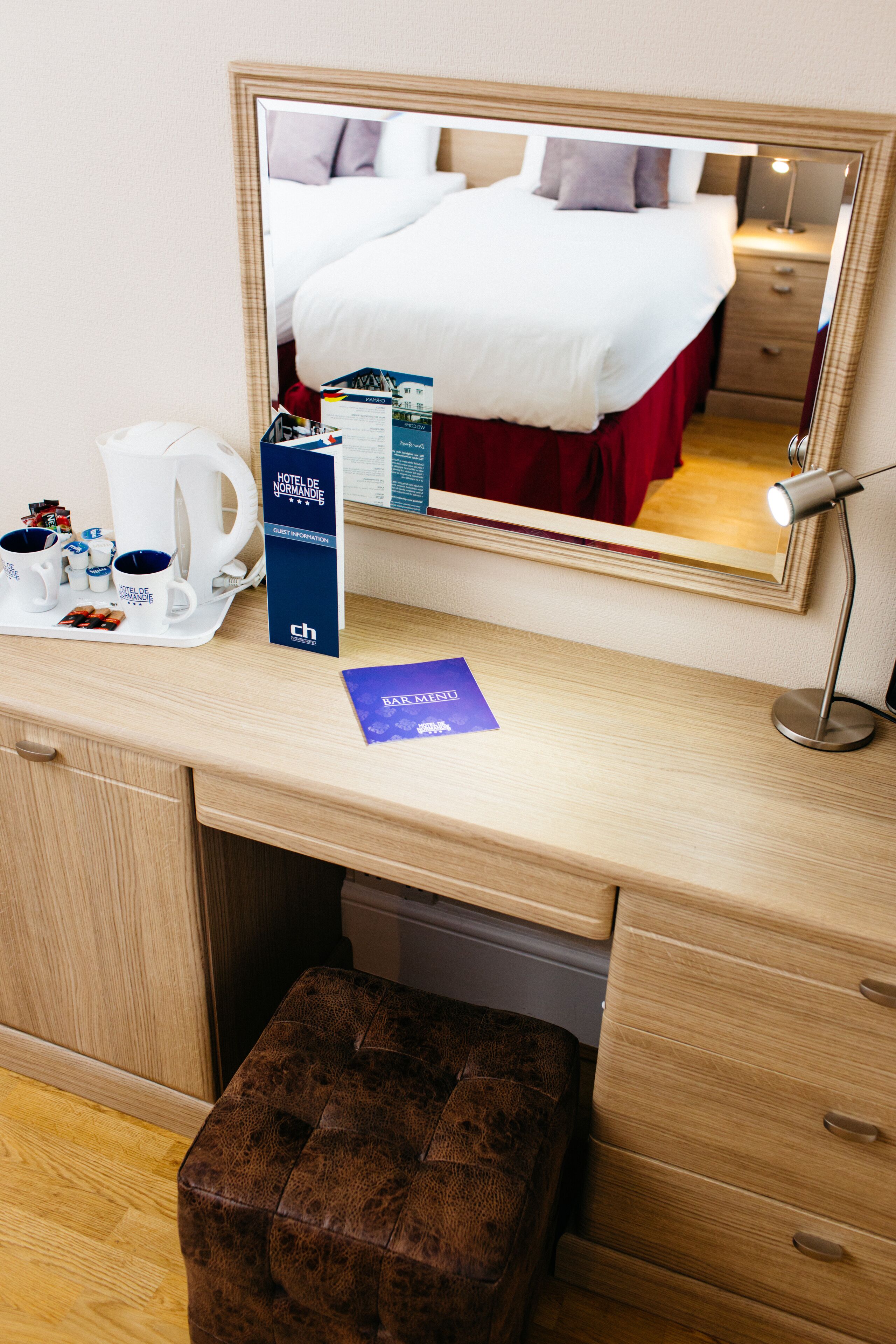 Une chambre d’hôtel avec un bureau en bois, un miroir, un lit avec une literie blanche, un pouf marron et une machine à café.
