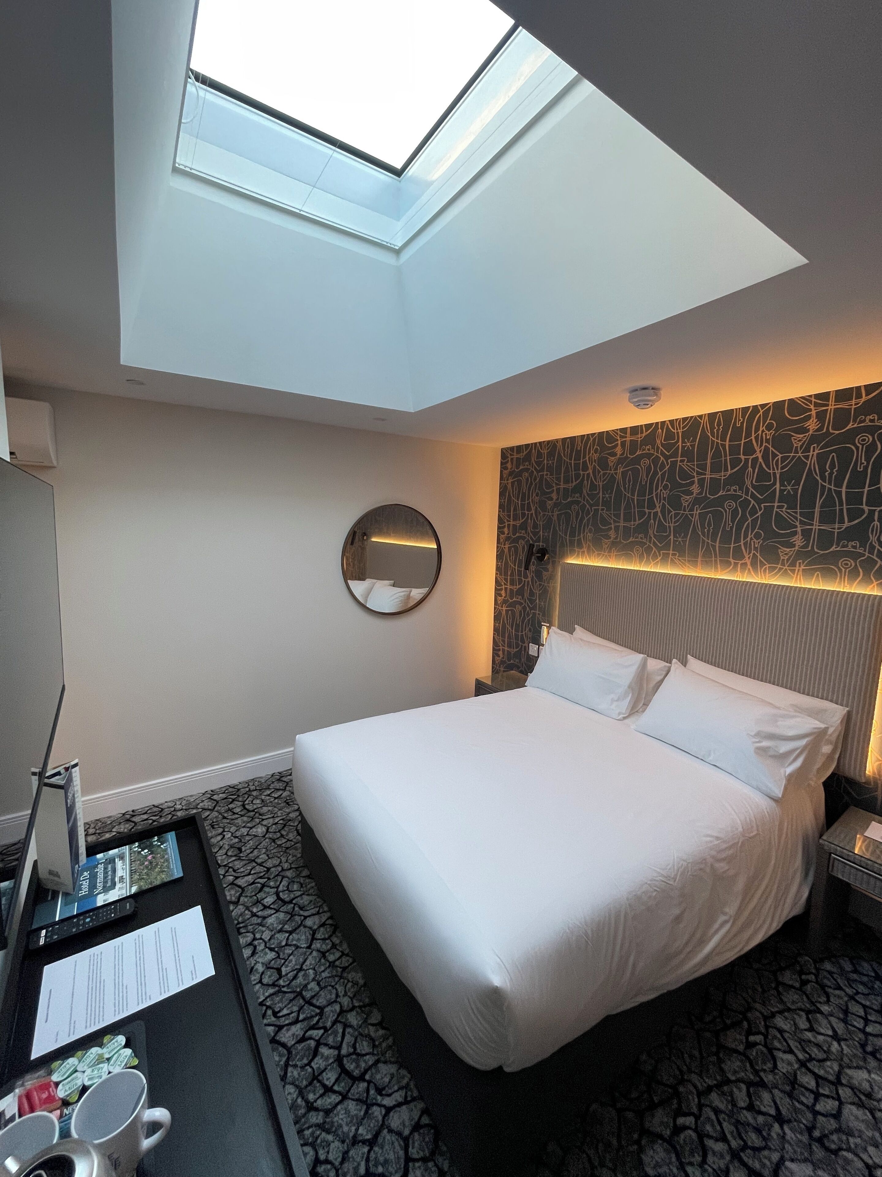 A hotel room with a skylight, a round mirror, a bed with white linens, a patterned wall, and a small table with a tray of items.
