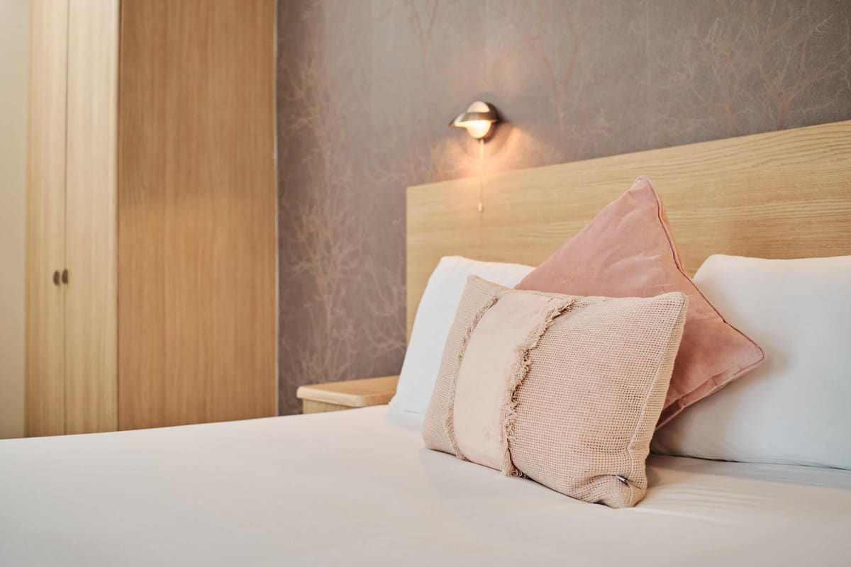 Una cama bien tendida con dos almohadas, un cabecero de madera y una mesita de noche con una lámpara.