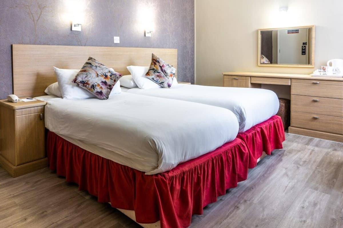 Una cama doble con falda roja, dos almohadas, una mesita de noche, un aparador de madera, un espejo y una lámpara de pared.