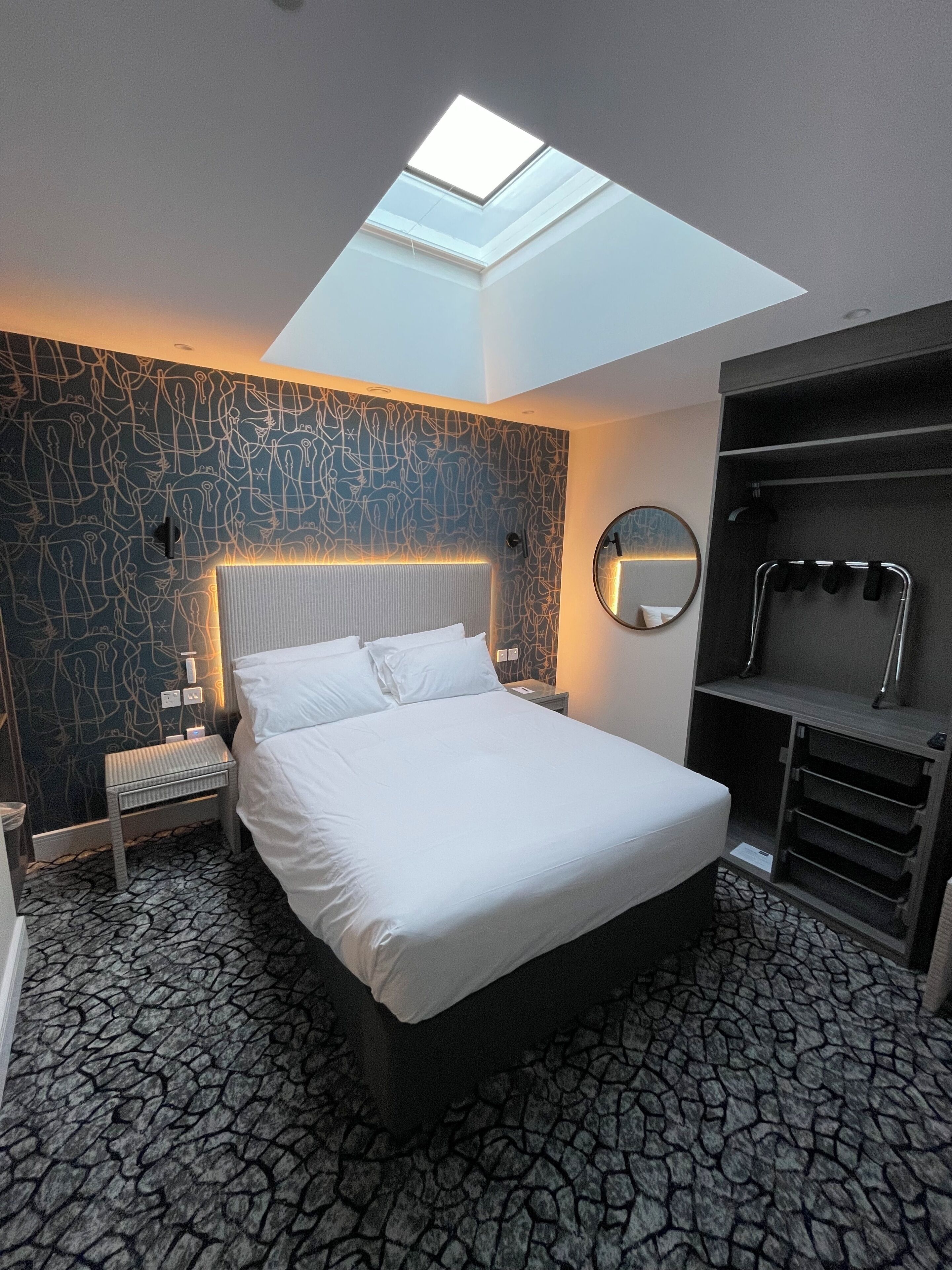 A modern hotel room with a skylight, a large bed, a bedside table, a mirror, and a built-in wardrobe.