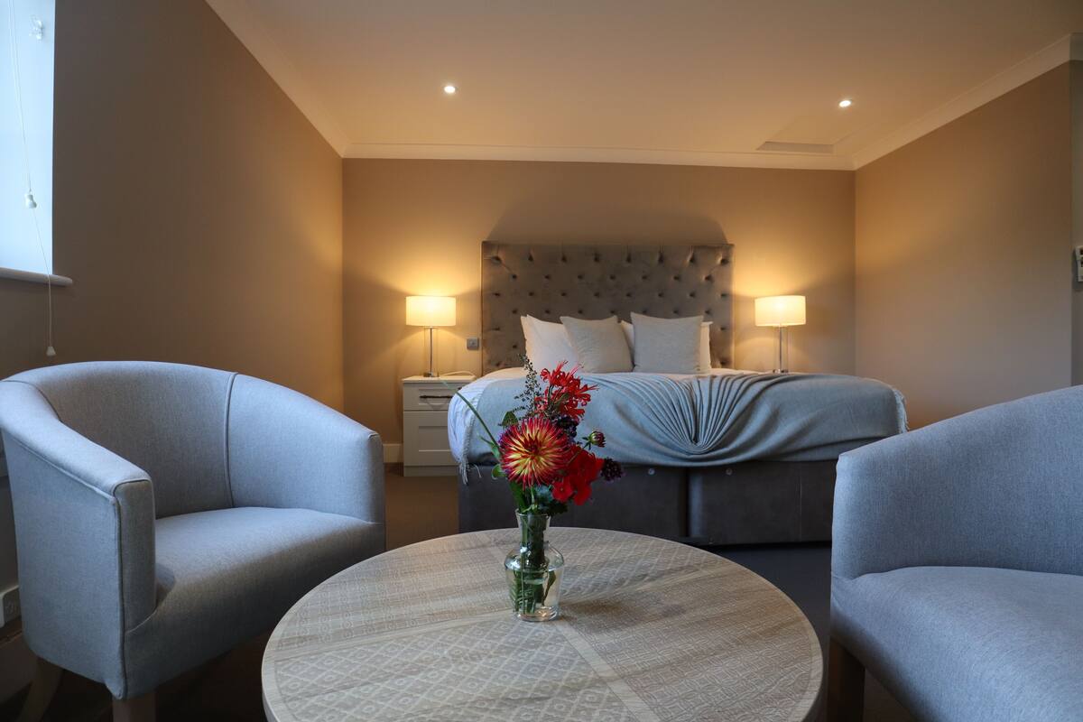 Habitación de hotel con cama, dos sillones, una mesa redonda con un florero y lámparas de noche.
