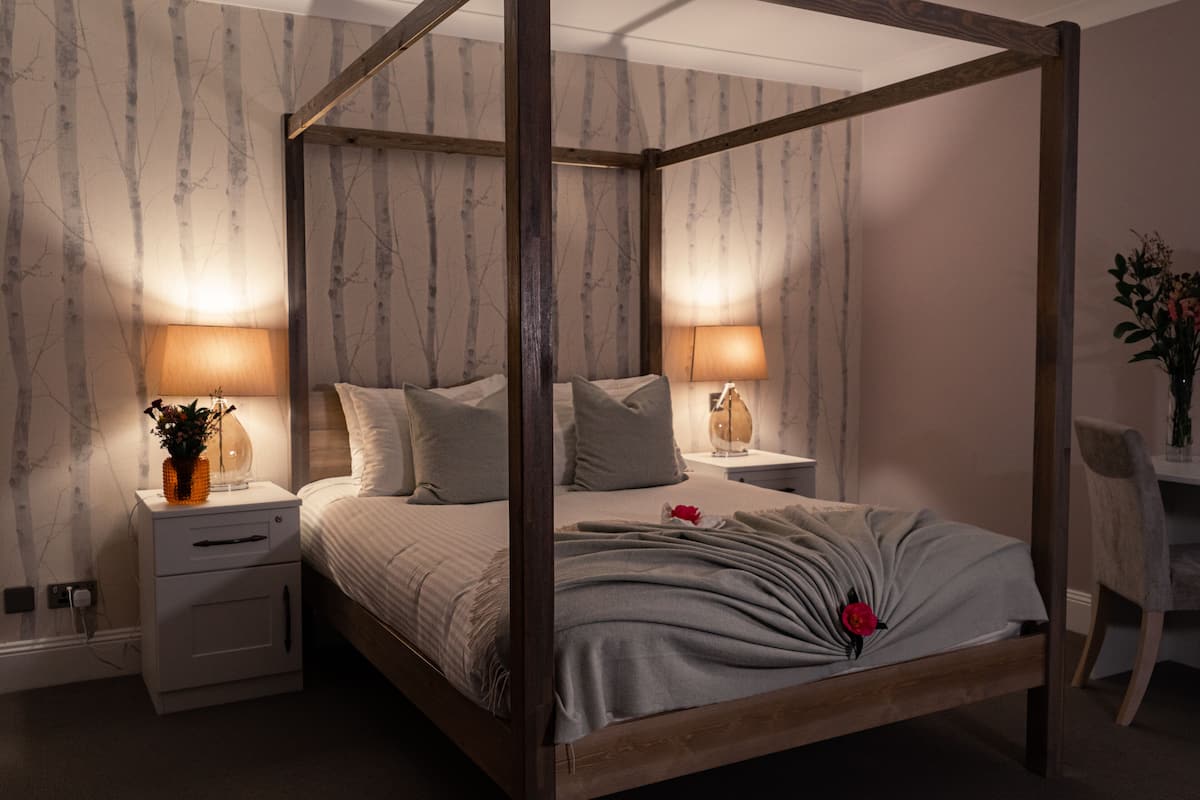 Una cama con dosel y estructura de madera, dos mesitas de noche con lámparas, una mesita auxiliar y una silla.