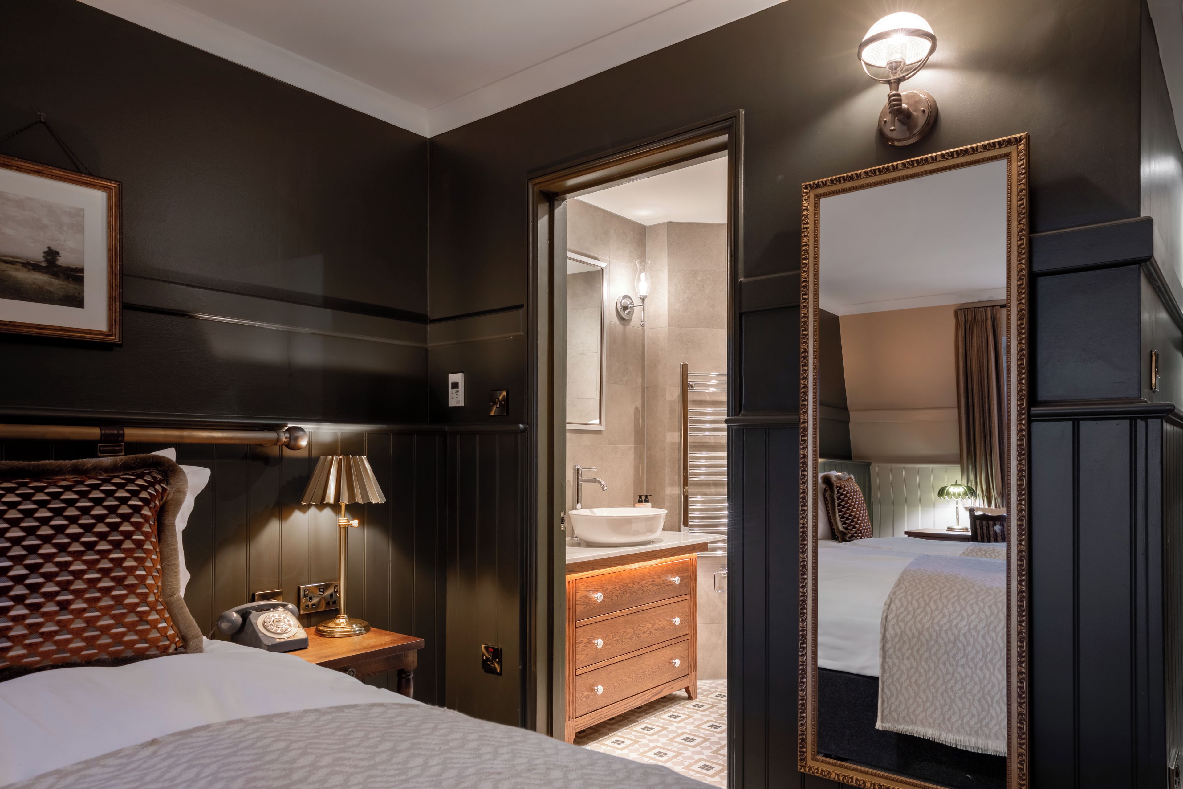 A hotel room with a bed, bedside table, lamp, dresser, mirror, and a bathroom visible through an open door.