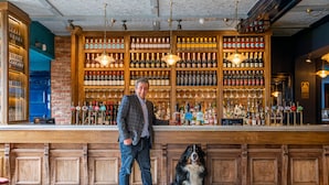 Un bar con estantes de madera llenos de botellas y vasos, un hombre de pie junto a un perro, y un suelo con diseño.