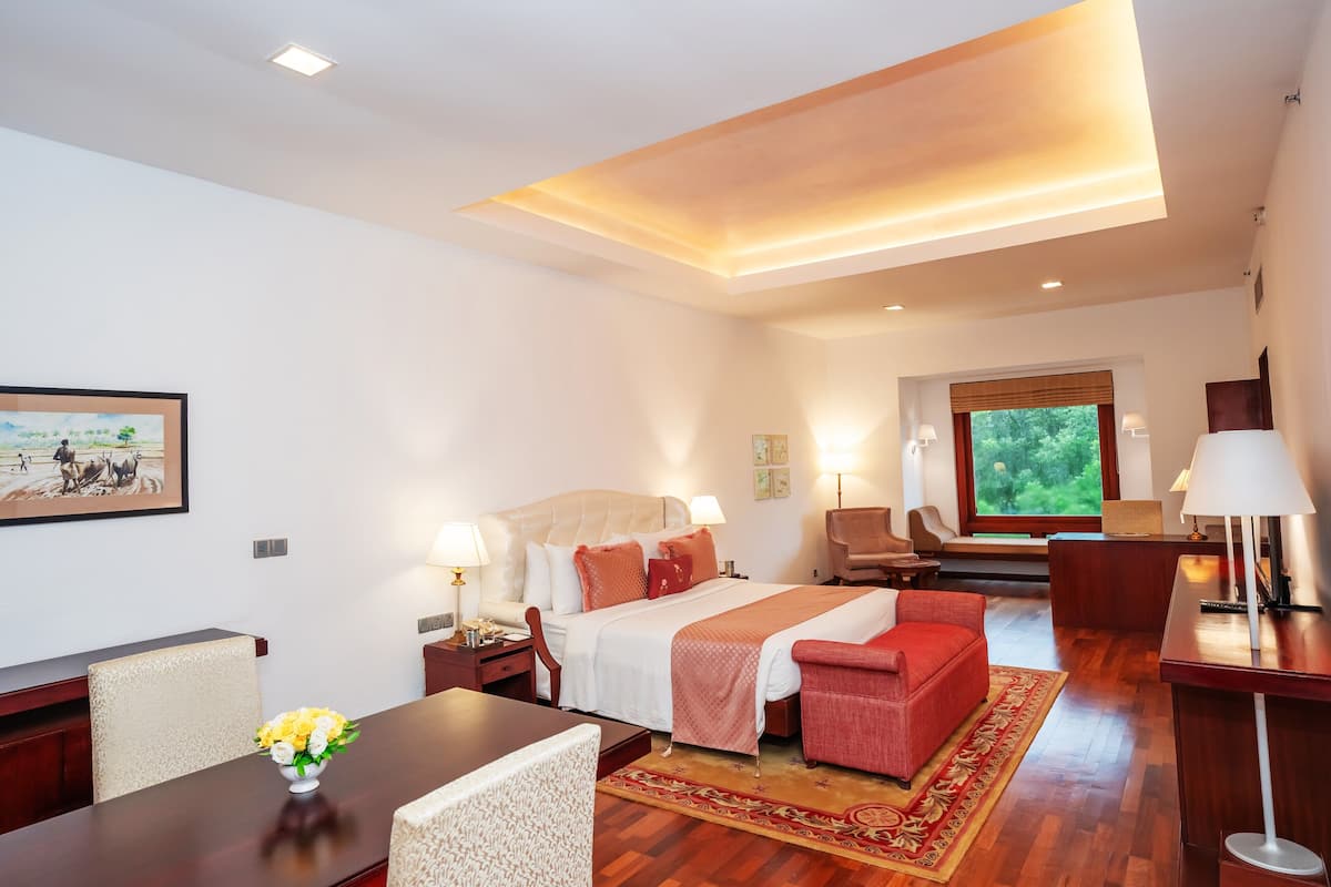 A hotel room with a bed, a desk, a chair, and a sofa. There is a window with a view of greenery outside.