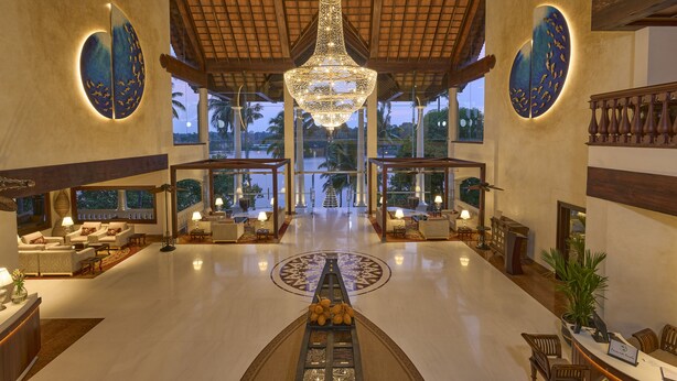 Un hall espacioso con una gran lámpara central, zonas de estar y amplias ventanas que ofrecen vistas a palmeras.