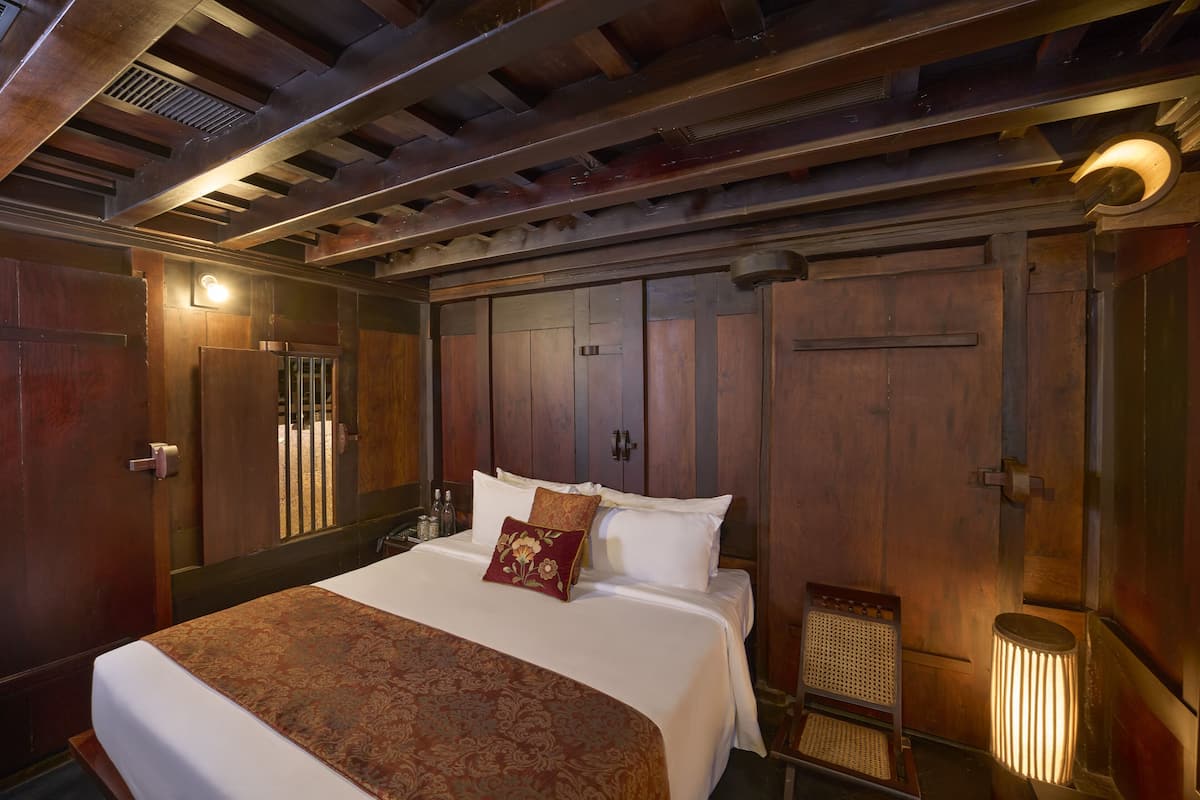 A large bed with a patterned bedspread and a single decorative pillow, wooden wardrobe, and a small chair with a woven seat.