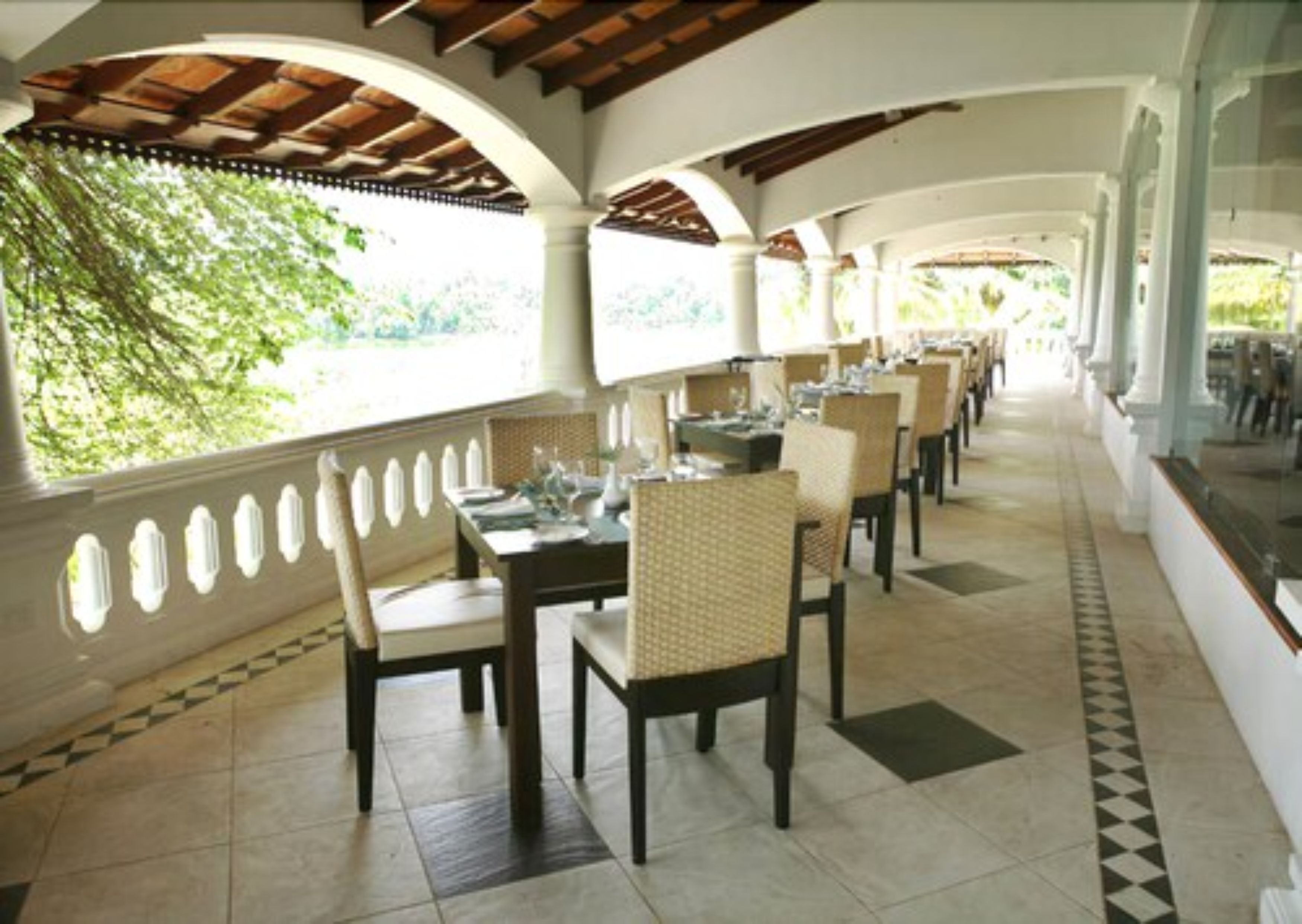Un área de comedor al aire libre techada, con mesas y sillas, ventanas amplias y piso de baldosas.