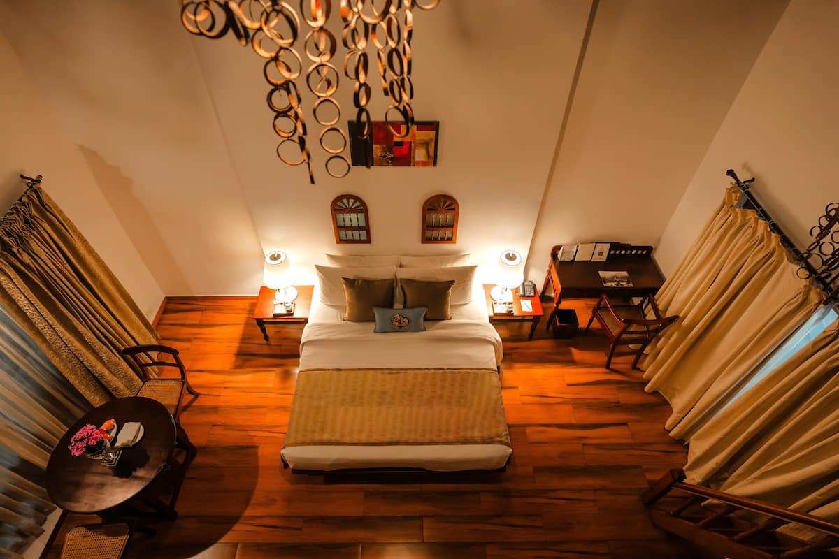 A hotel room with a large bed, wooden flooring, curtains, a chandelier, and two bedside tables with lamps.