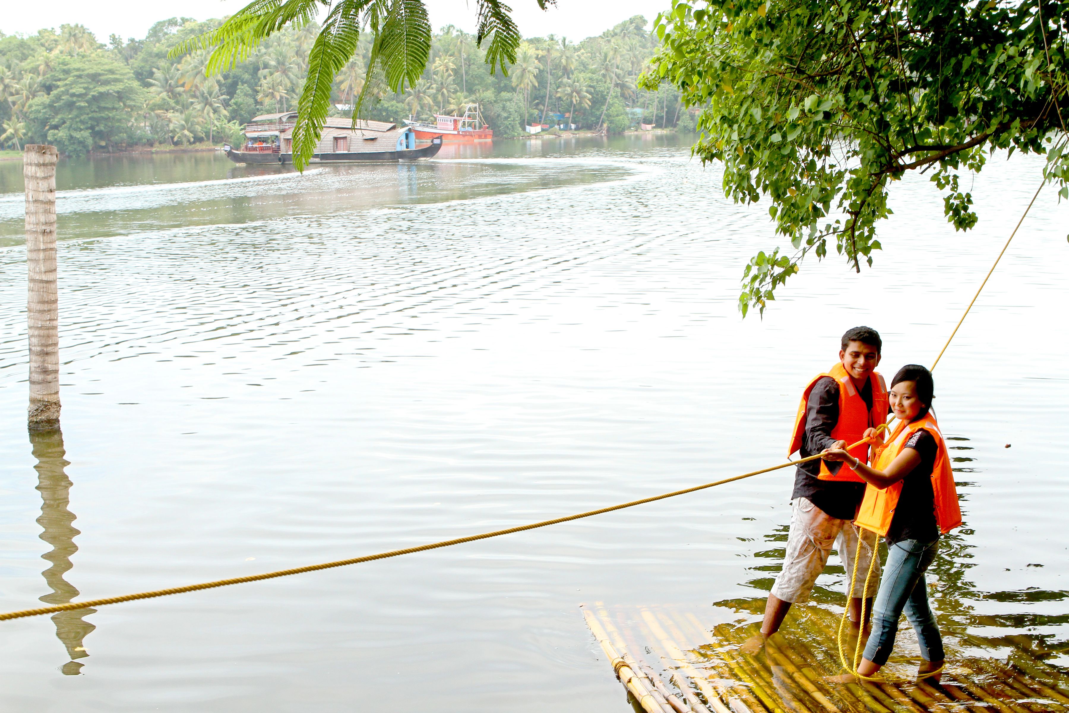 Two people on a floating platform in a calm body of water, with boats and greenery in the background.