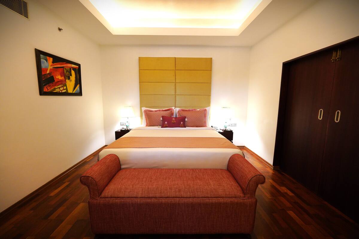 A hotel room with a large bed, a wooden floor, a bench, a painting on the wall, and a wardrobe.