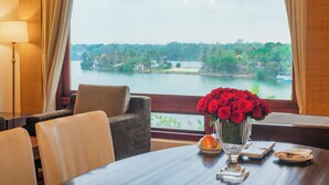 Un comedor con una mesa dispuesta para dos, un jarrón de rosas rojas y una vista de un lago con edificios y árboles al fondo.