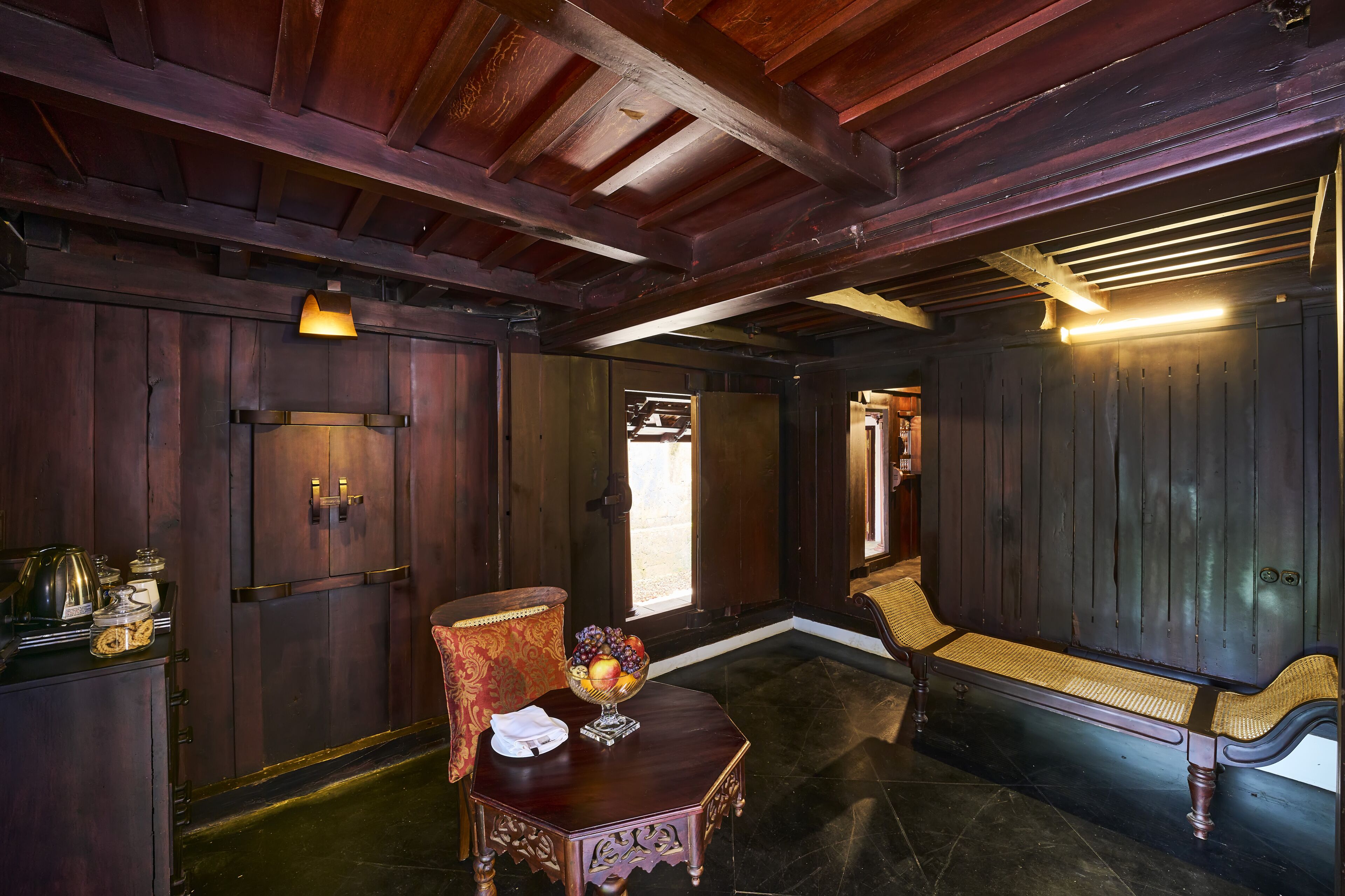 A wooden-paneled room with a bench, a small table with a fruit bowl, and a lamp.