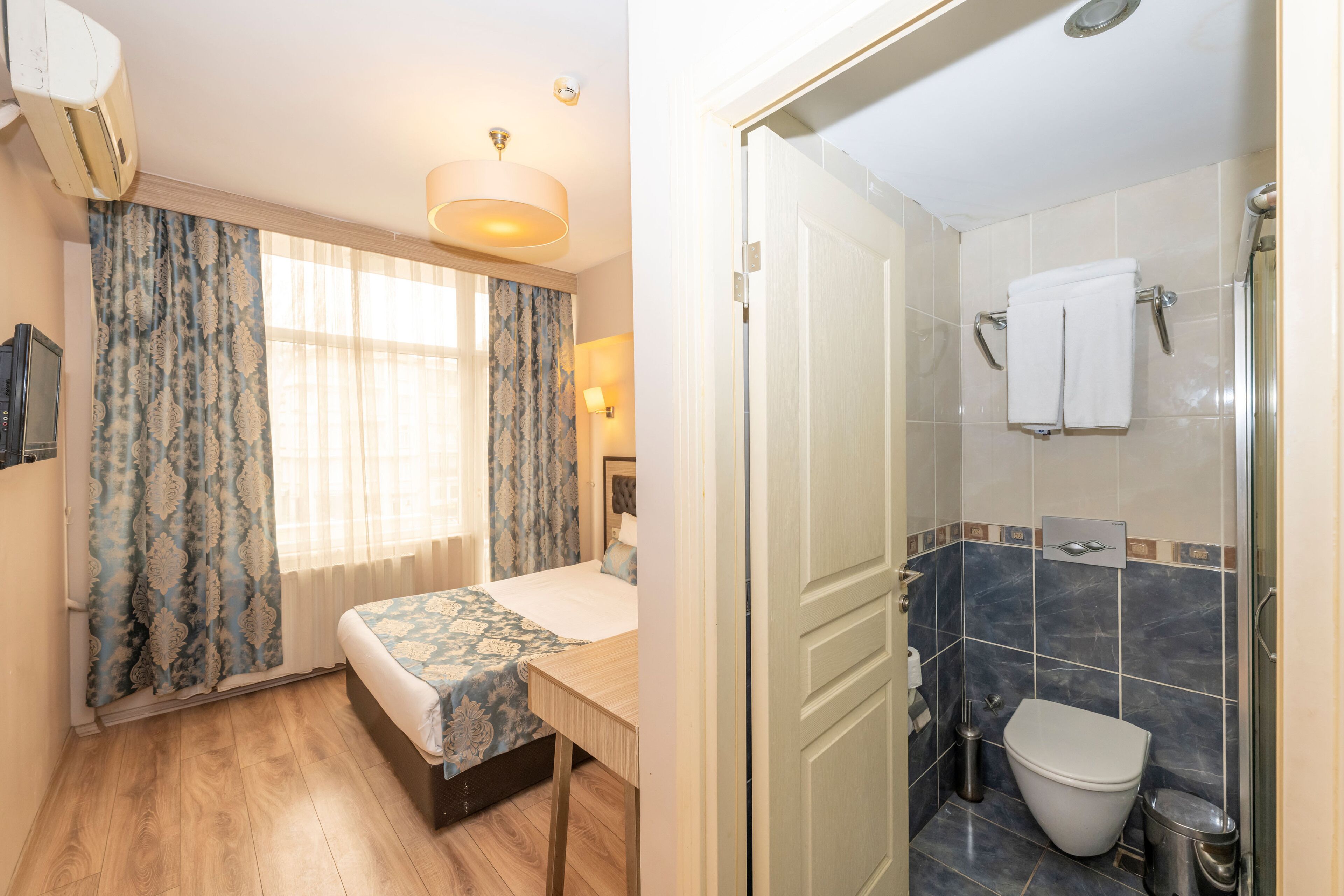 A bedroom with a bed, a bedside table, and a window with curtains. The bathroom includes a toilet, a sink, and a shower area.