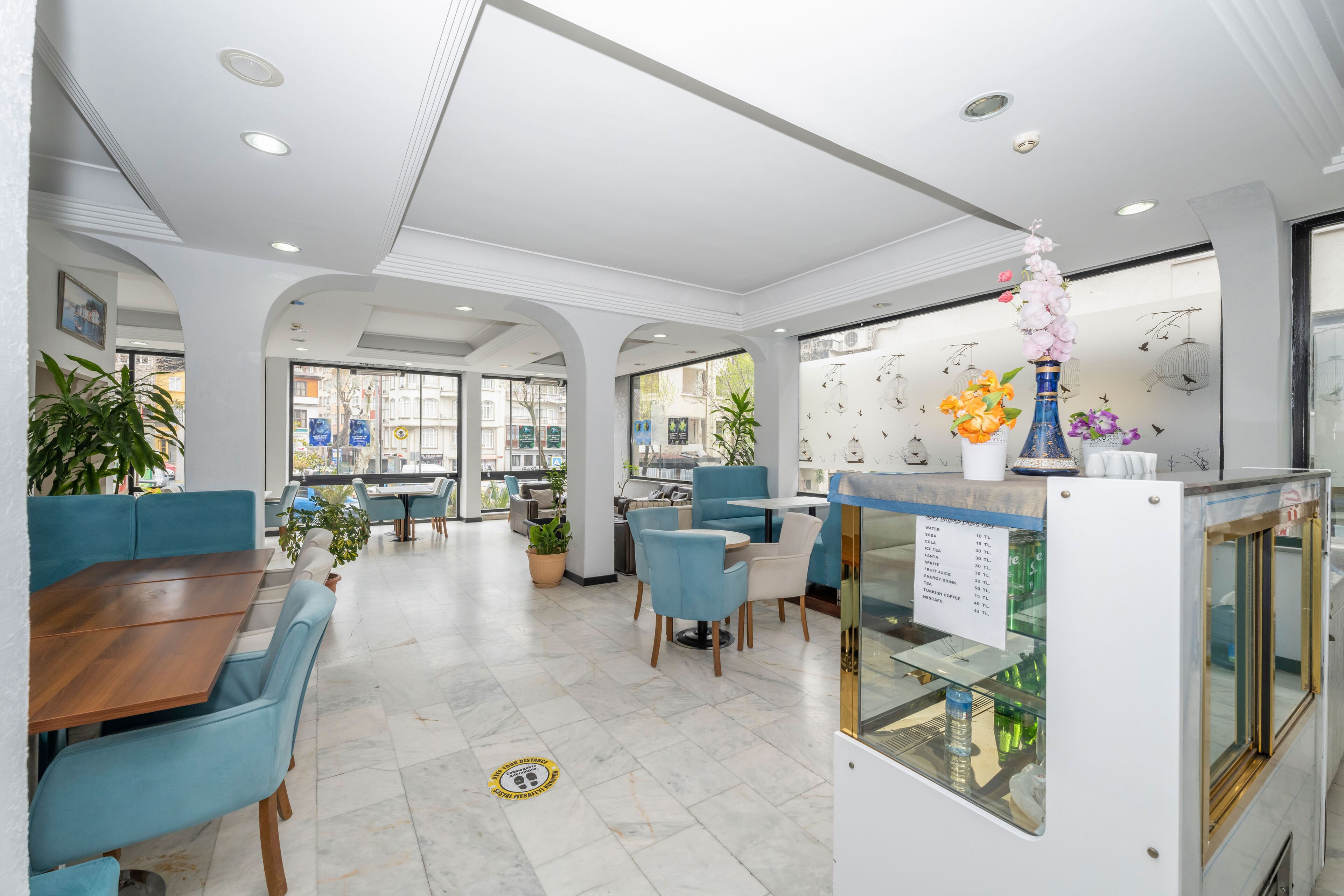 A modern dining area with blue upholstered chairs and a wooden table, a reception desk with a menu, and potted plants.