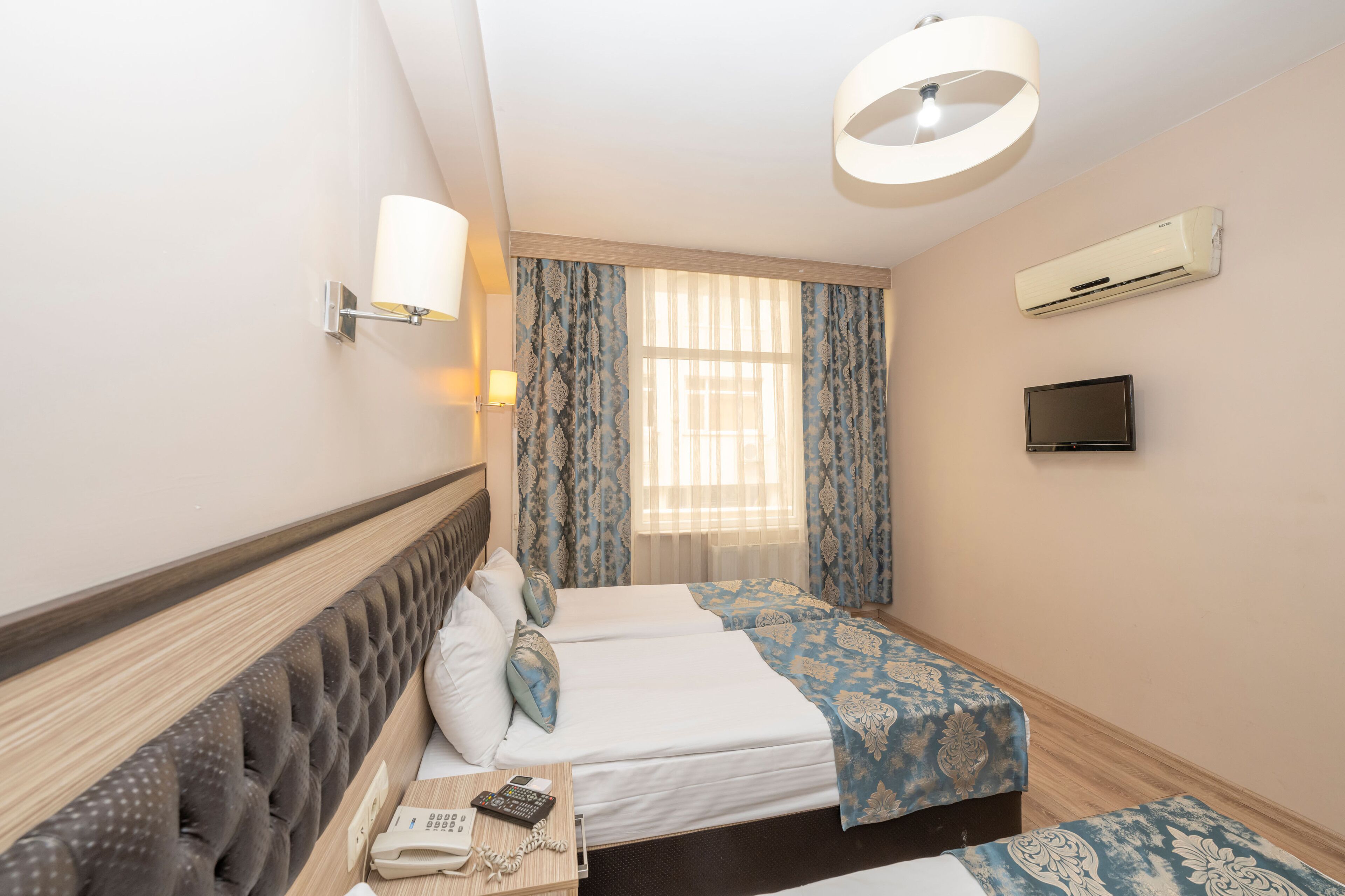 A hotel room with two beds, a flat-screen TV, an air conditioning unit, a bedside lamp, and a window with curtains.