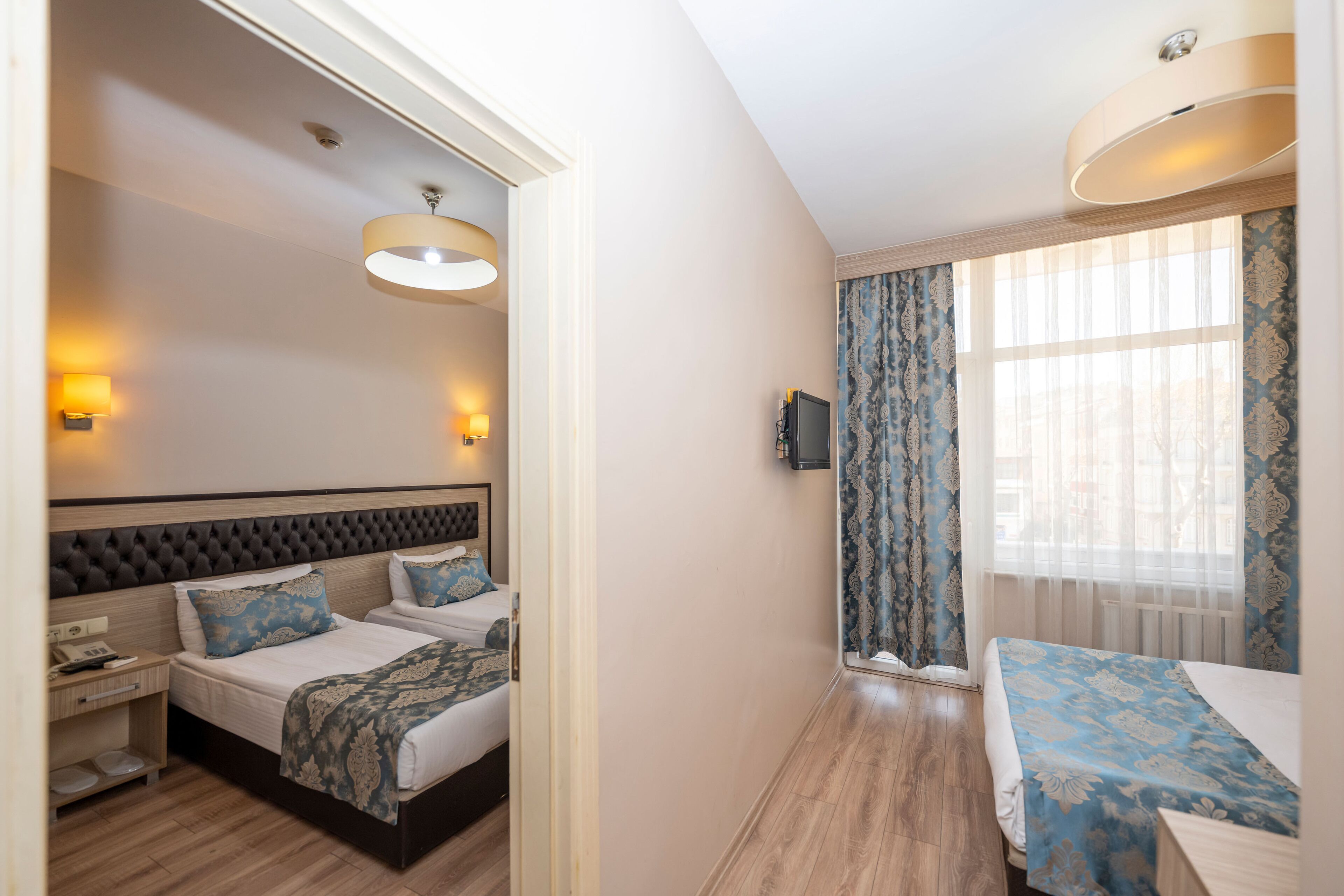 A hotel room with two beds, a wooden floor, a large window with curtains, and a wall-mounted TV.