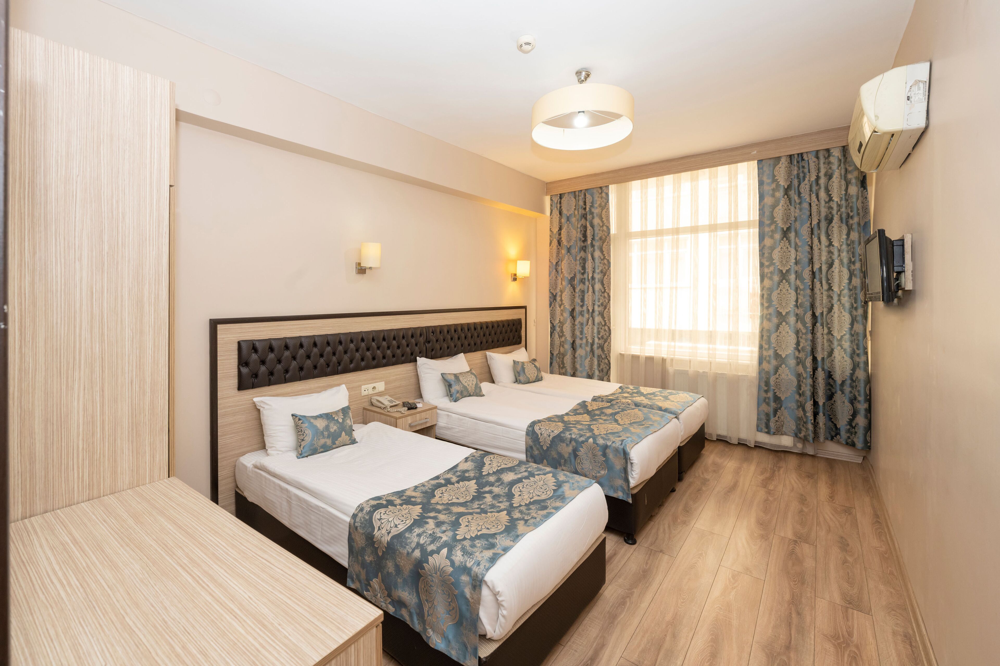 A hotel room with two beds, a desk, a wardrobe, a ceiling light, and a window with curtains.