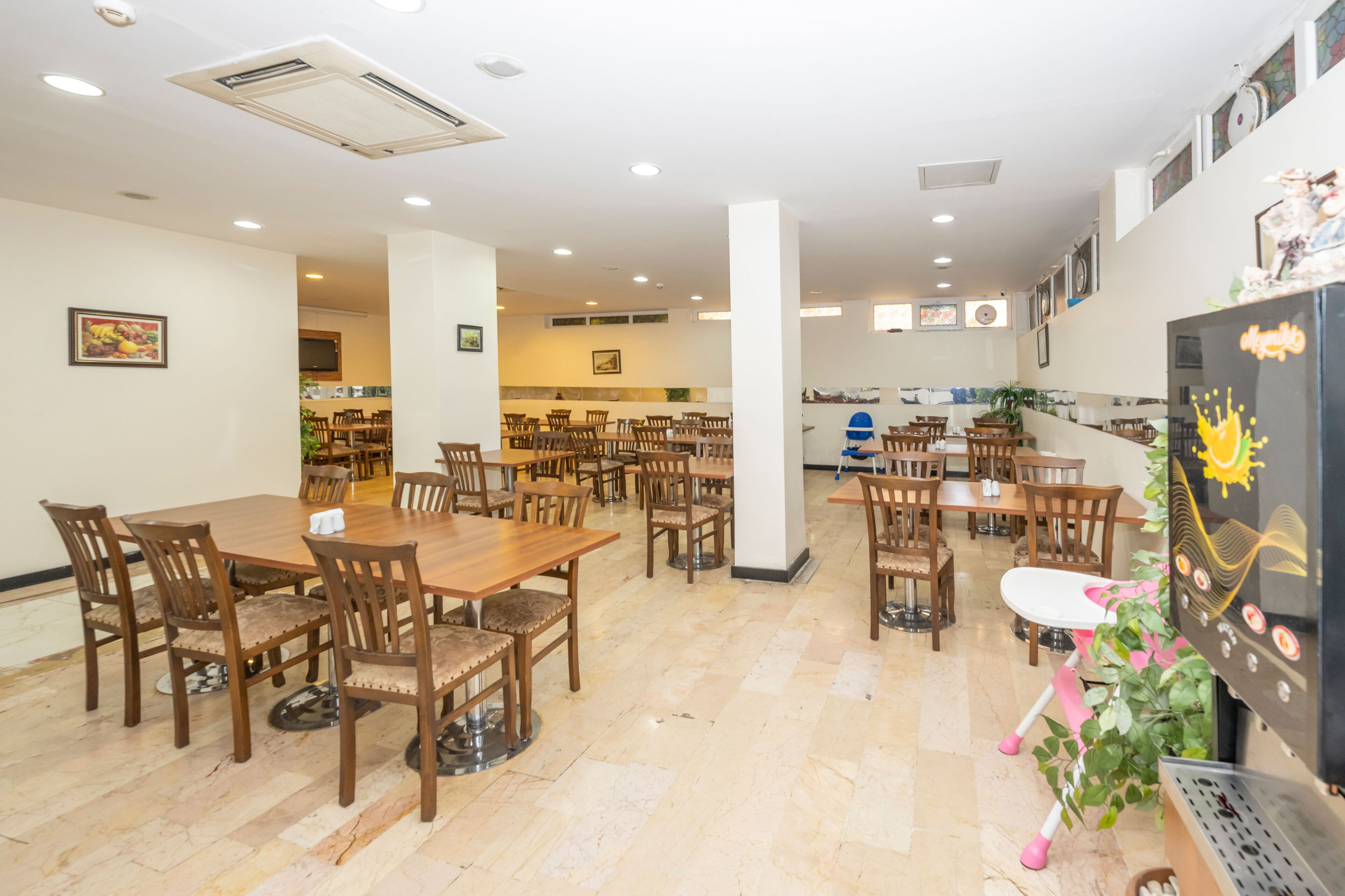 A spacious dining area with multiple wooden tables and chairs, a kitchen in the background, and a refrigerator with decorative magnets.