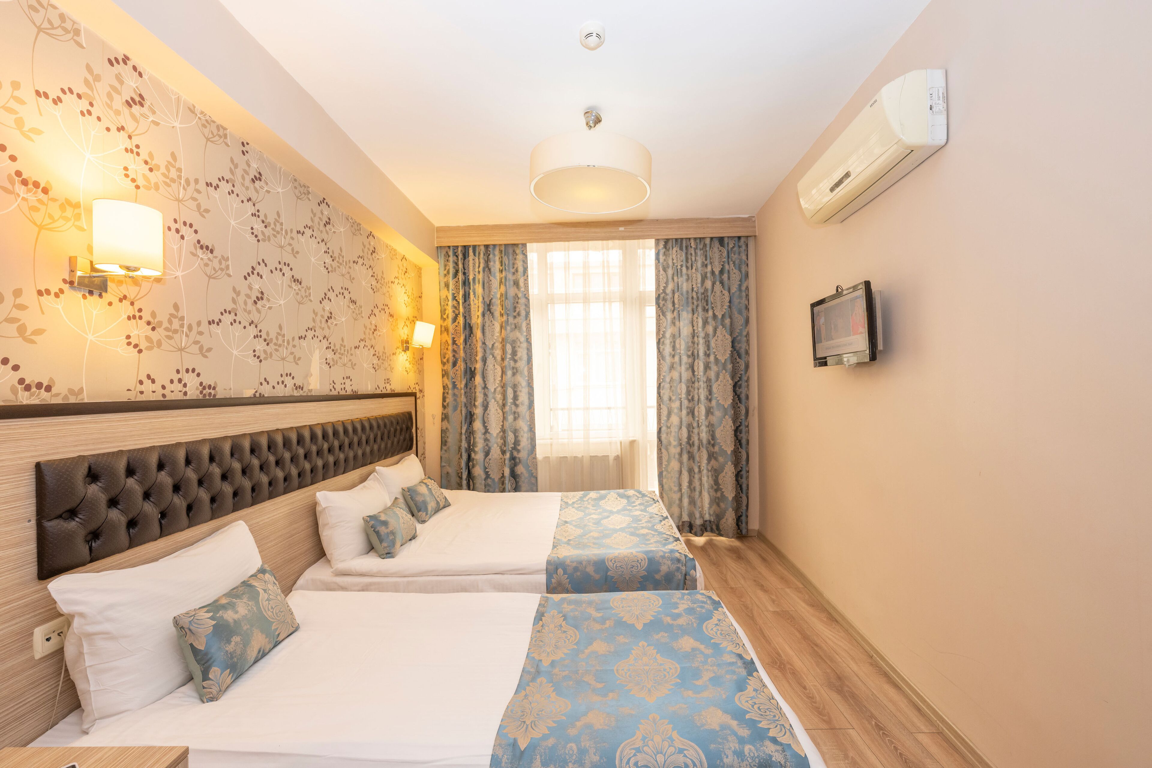 A hotel room with two beds, a large window with curtains, a wall-mounted TV, and an air conditioning unit.