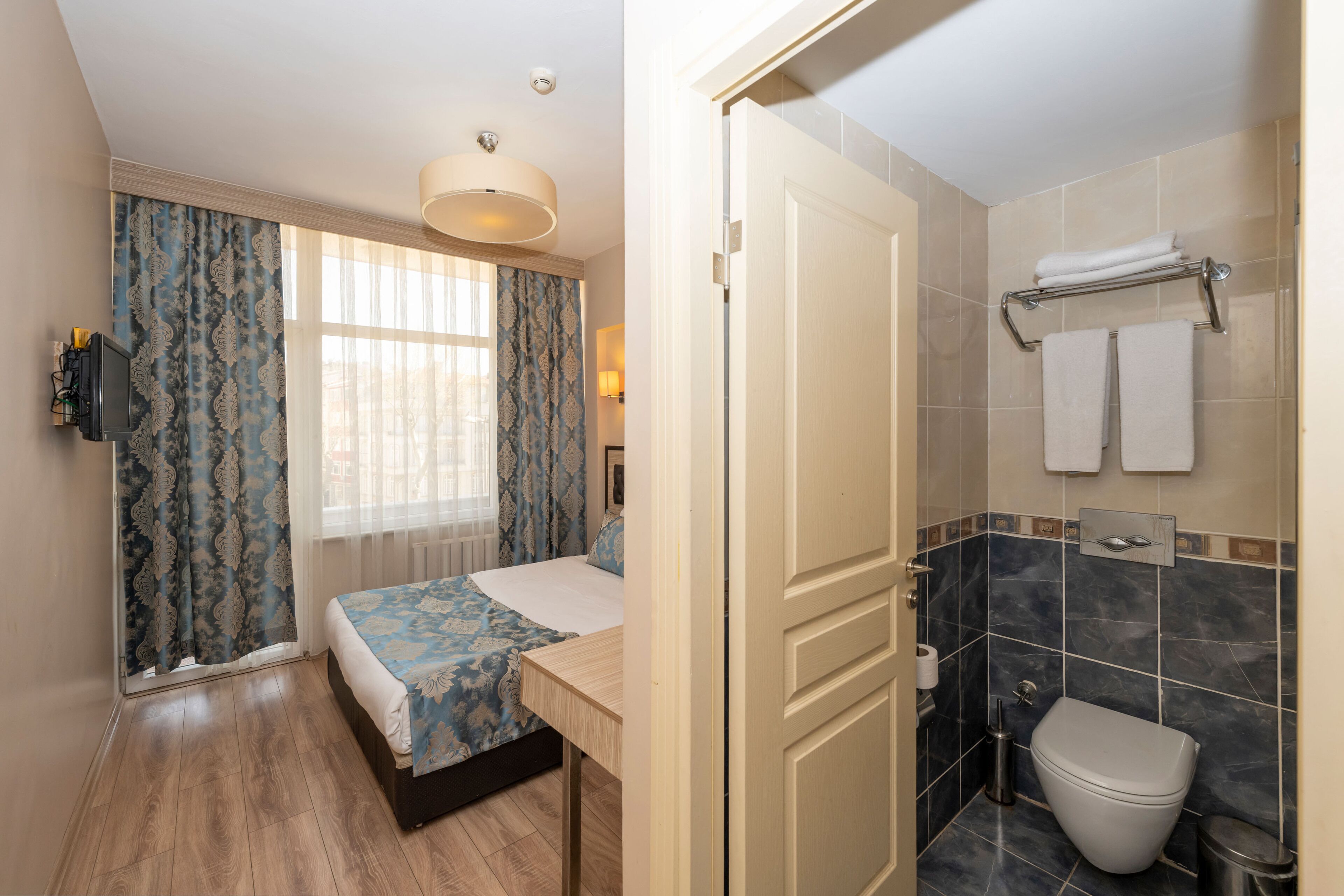 A bedroom with a bed, a bedside table, a window with curtains, a bathroom with a toilet, a towel rack, and a shower area.
