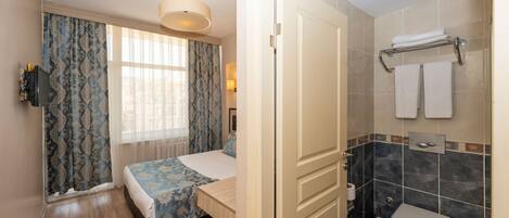 A bedroom with a bed, a bedside table, a window with curtains, a bathroom with a toilet, a towel rack, and a shower area.