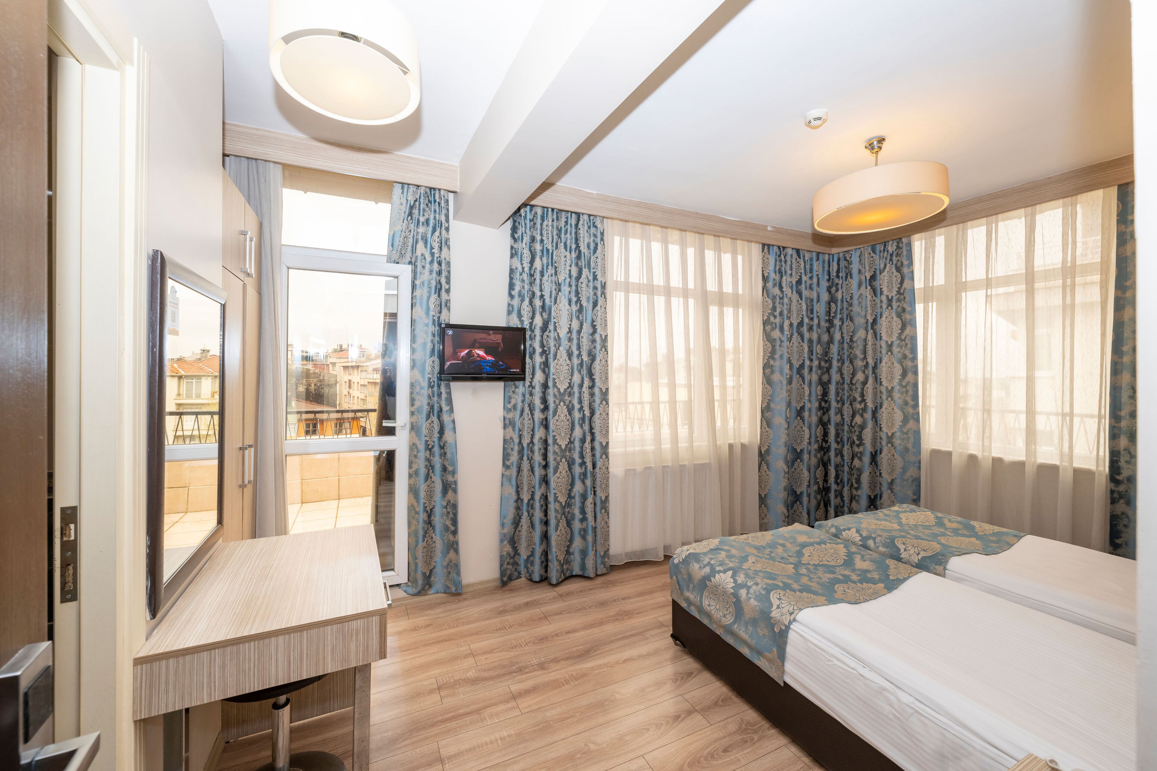 A hotel room with a bed, a desk, a television, and large windows with curtains.