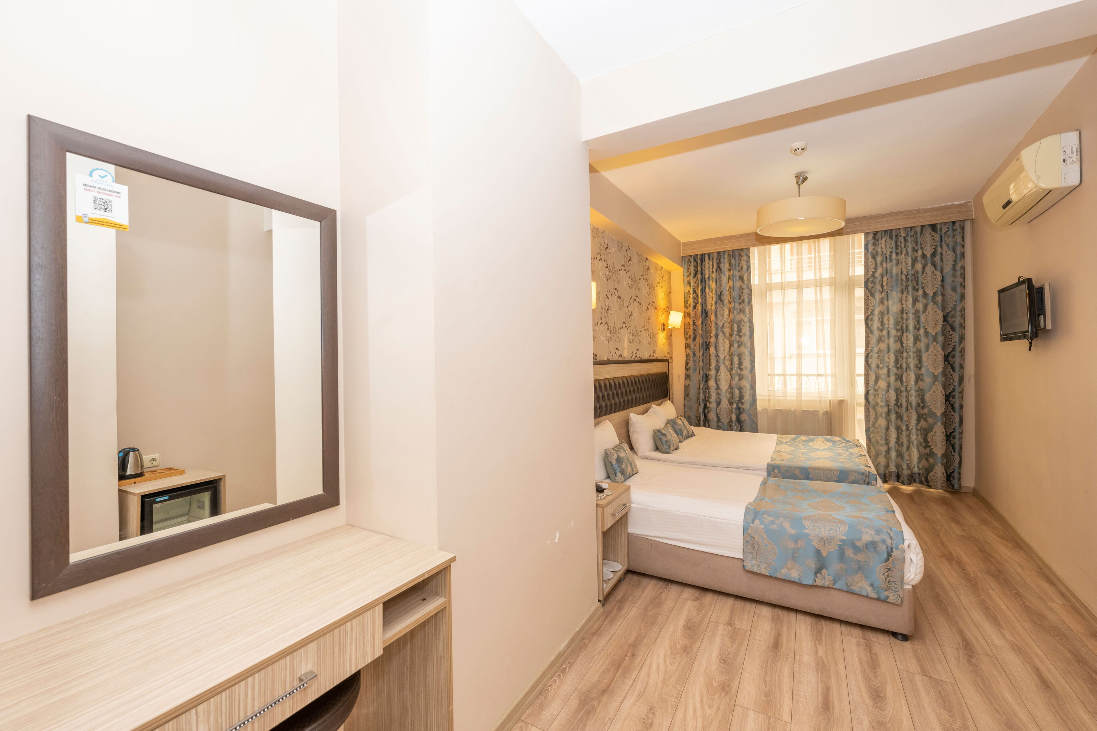 A hotel room with a large mirror, a bed with blue bedding, a bedside table, and a small desk with a lamp.