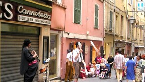 A street scene with a group of people, some sitting and others standing, in front of a building with a closed shutter. - Via Palazzo Sanremo (Sanremo)