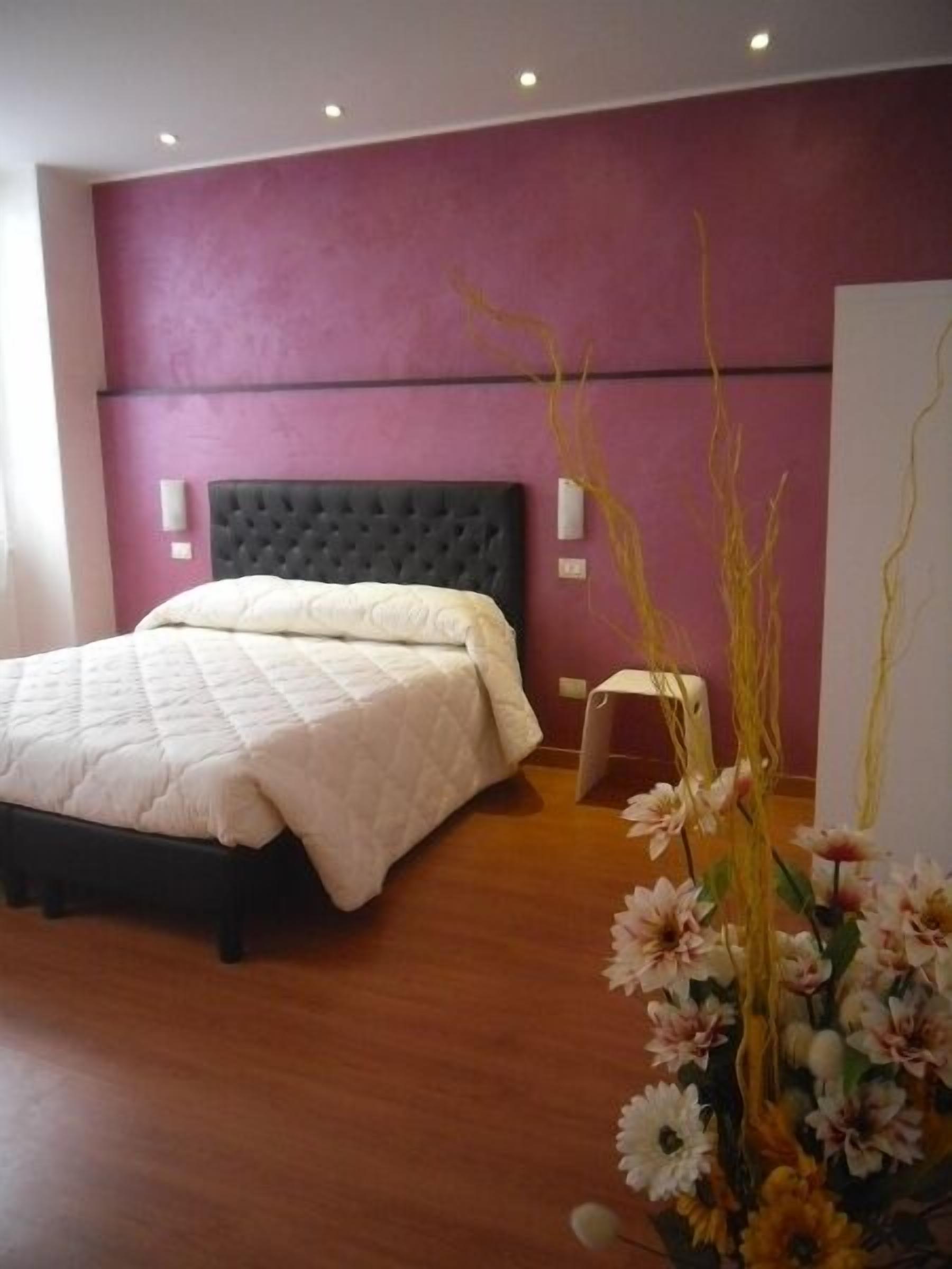 A bedroom with a bed, a nightstand, a vase with flowers, and a wall with a light switch.