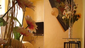 A room with a wooden floor, a flat-screen TV mounted on the wall, a large white vase with flowers, and a traditional fan. - Via Palazzo Sanremo (Sanremo)