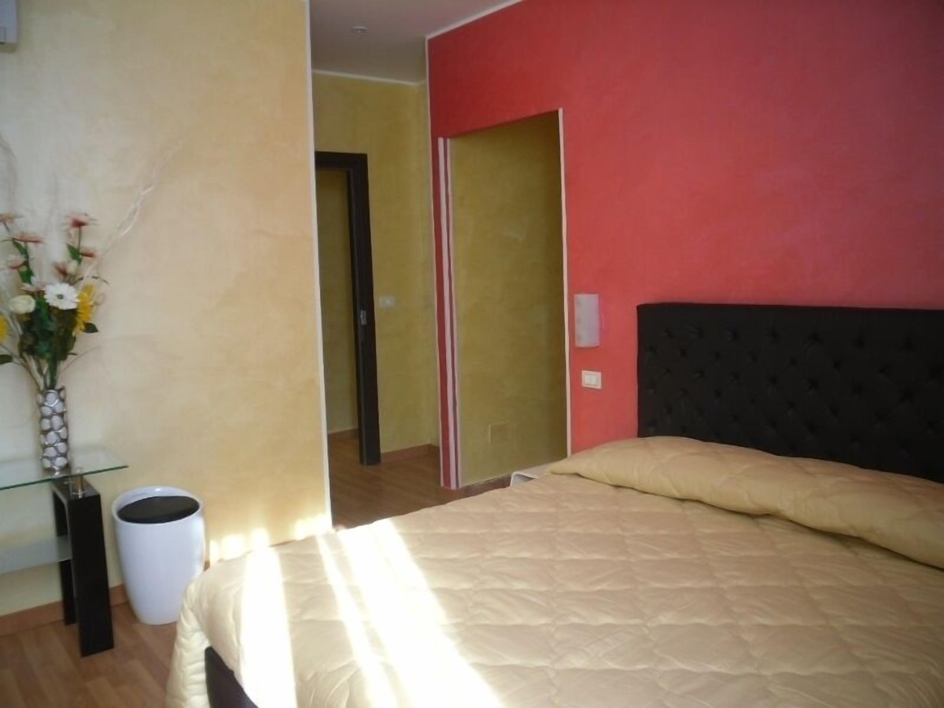 A bedroom with a bed, a vase with flowers, a trash bin, and a door leading to another room.