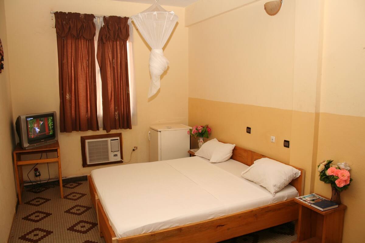 A room with a bed, a television on a stand, a small refrigerator, and a bedside table with a vase of flowers.