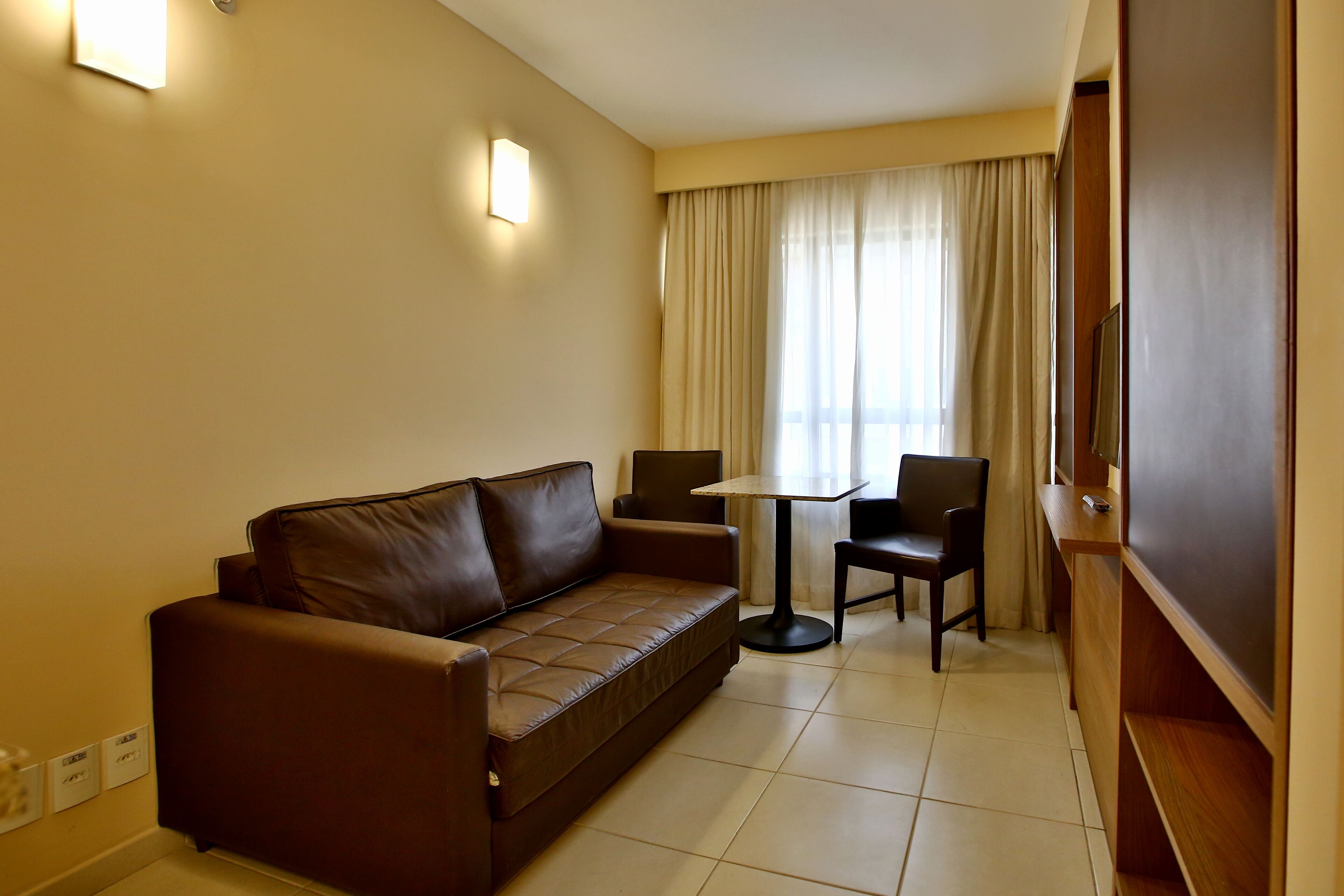 Une chambre d’hôtel avec un canapé en cuir marron, une petite table ronde, deux chaises, un téléviseur et une fenêtre avec des rideaux.