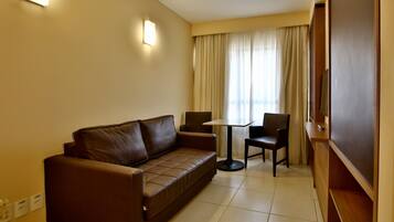 A hotel room with a brown leather sofa, a small round table, two chairs, a TV, and a window with curtains.