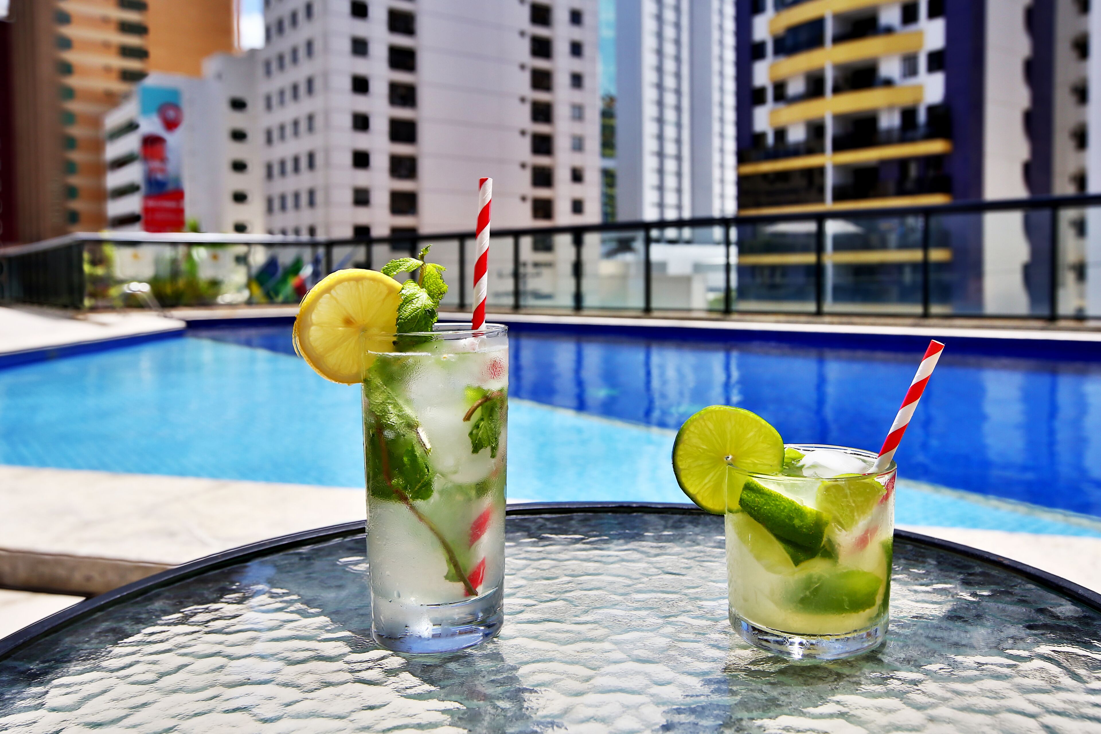 Dos vasos de una bebida de lima con hojas de menta y una rodaja de lima sobre una mesa de vidrio junto a una piscina, con un paisaje urbano al fondo.
