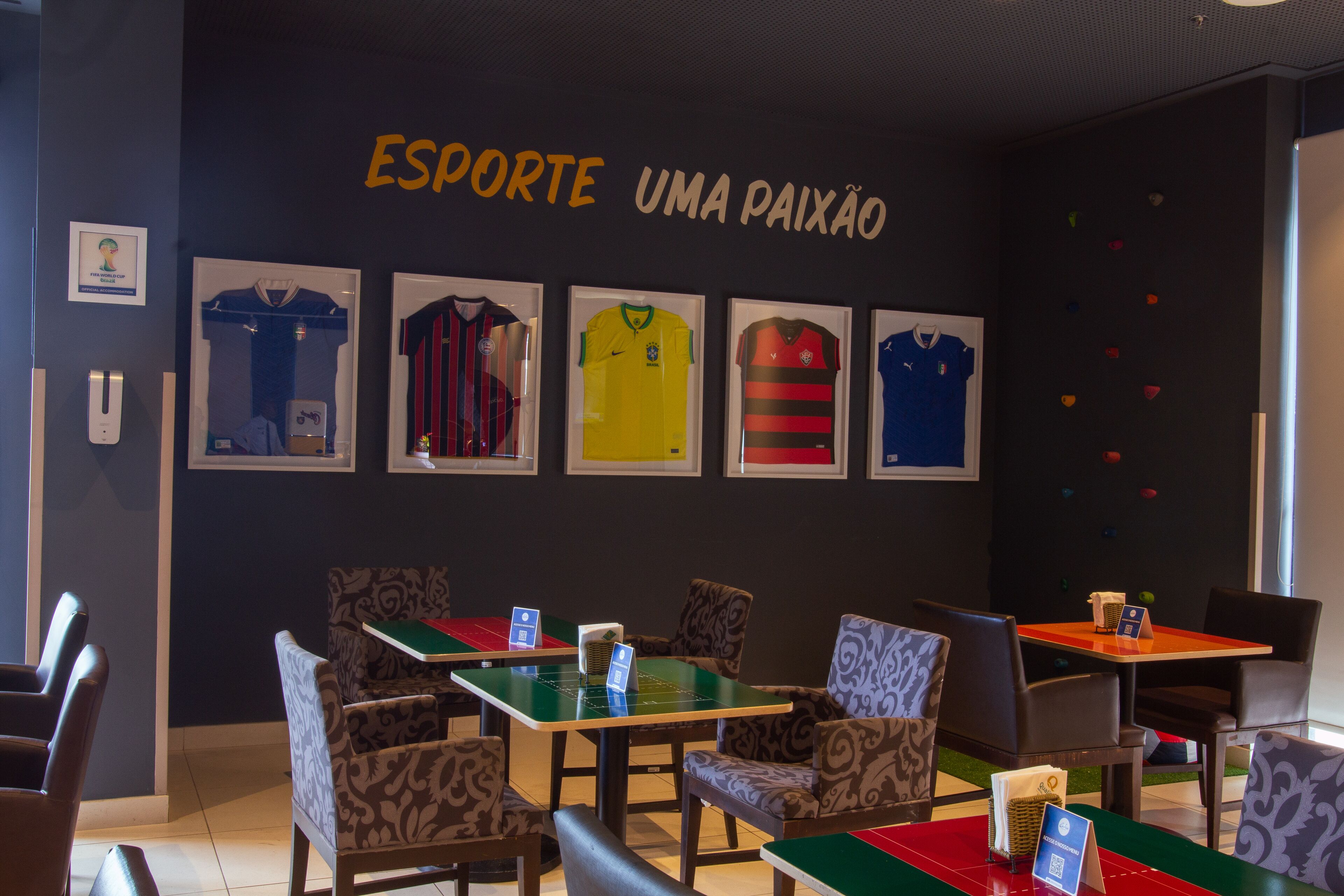 Uma área de refeições com mesas e cadeiras, parede com camisas emolduradas e um letreiro com a frase "ESPORTE UMA PAIXÃO".