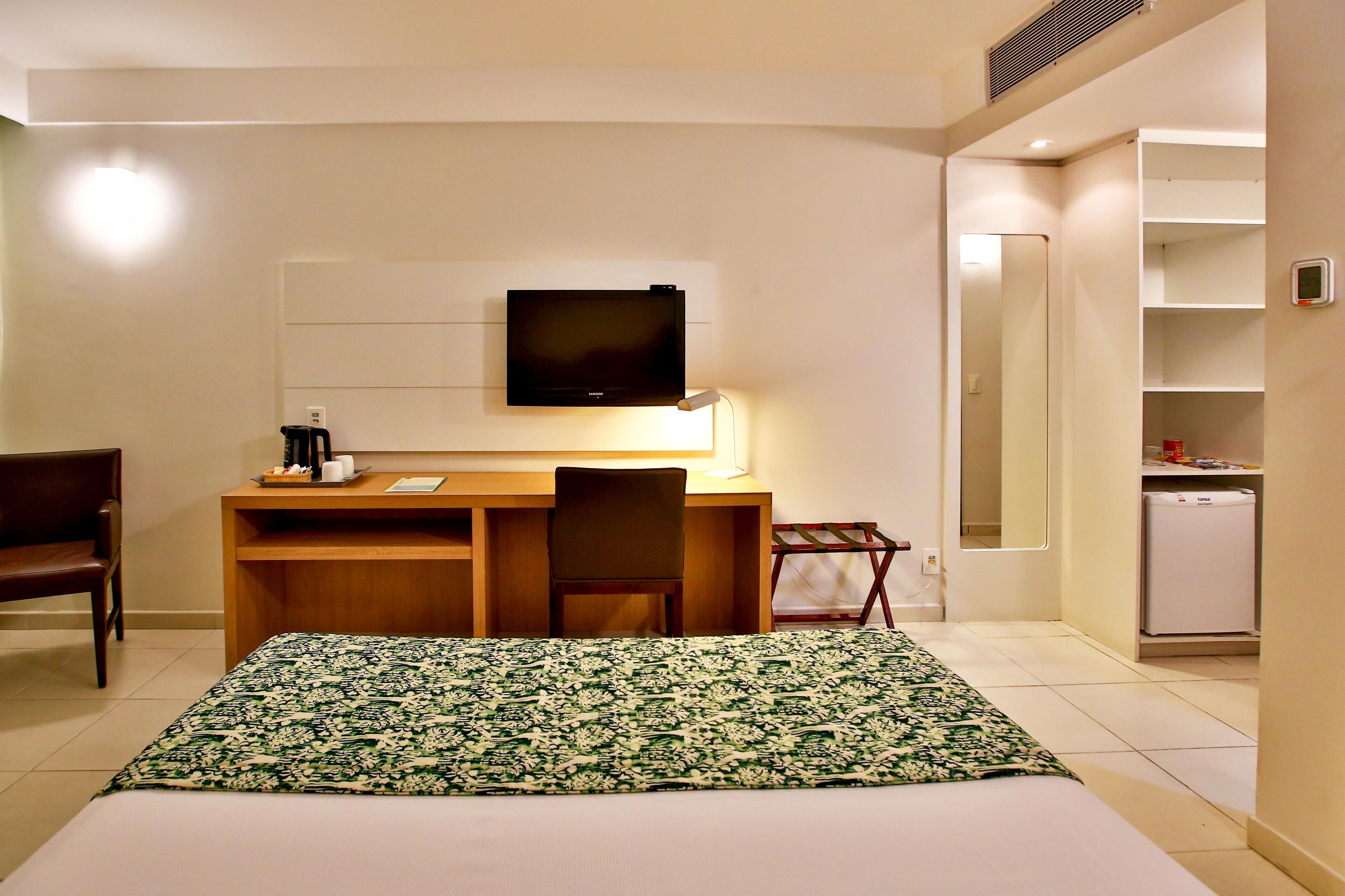 Ein Hotelzimmer mit Bett, Schreibtisch mit Stuhl, einem an der Wand befestigten Fernseher und einem kleinen Kühlschrank.